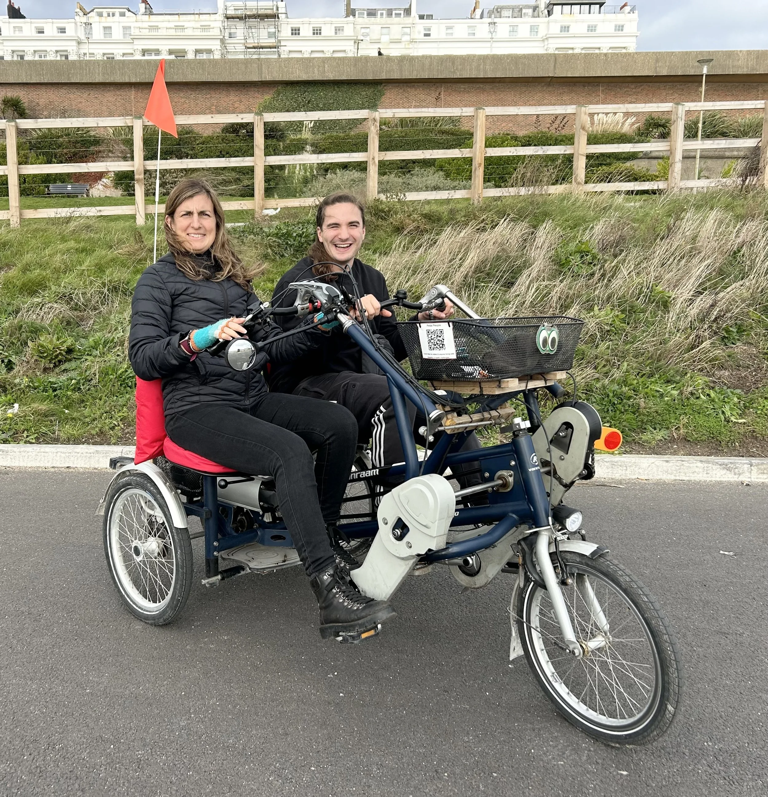 Two participants sit on a side-by-side tandem and they are smiling at the camera. Behind them is a grassy bank and fence. Buildings can be seen in the background.