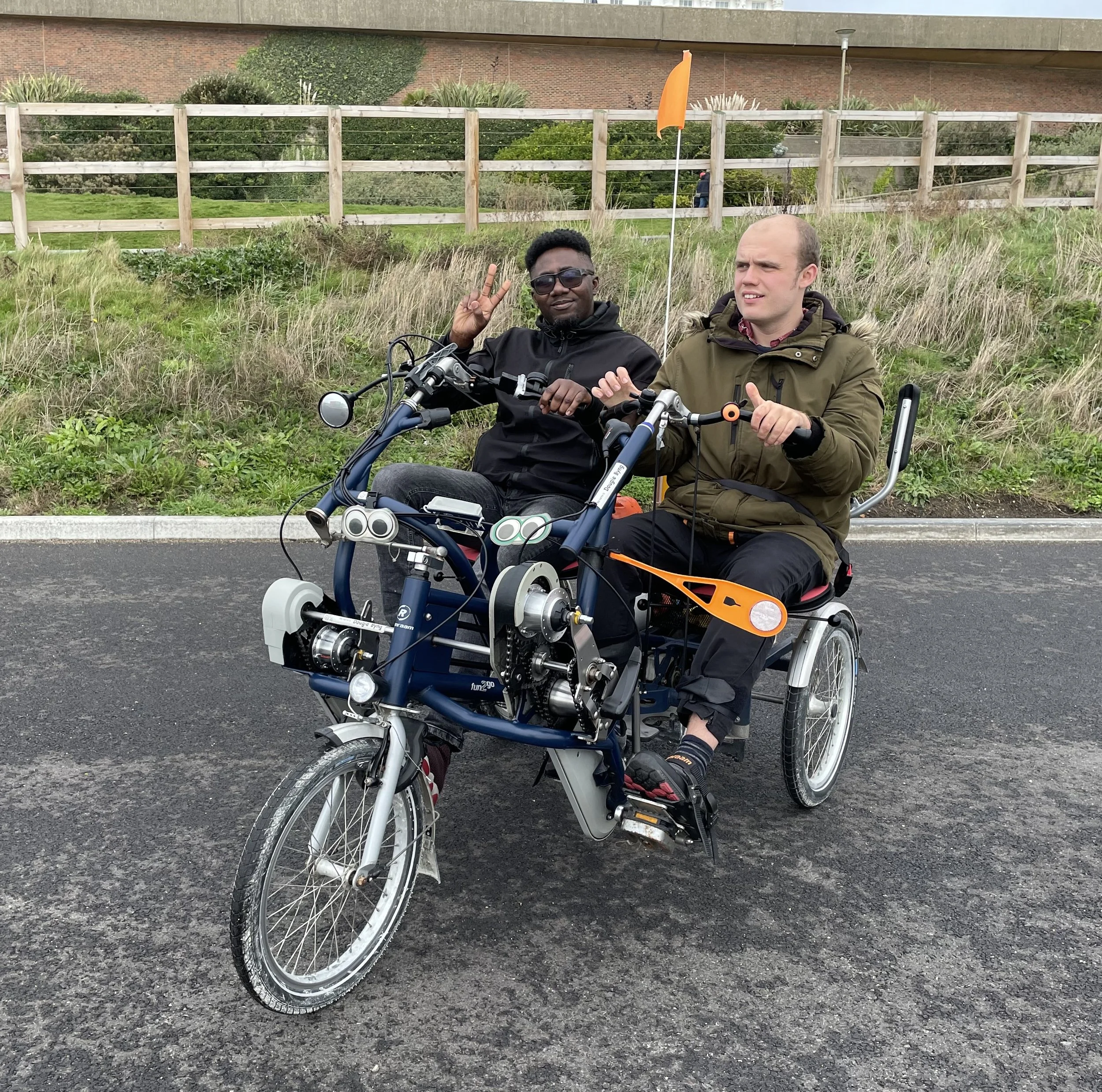 Two participants are sitting on a side-by-side tandem. One of them is doing a 'peace' sign with their hand. They are parked in front of a grassy bank and wooden fence. 