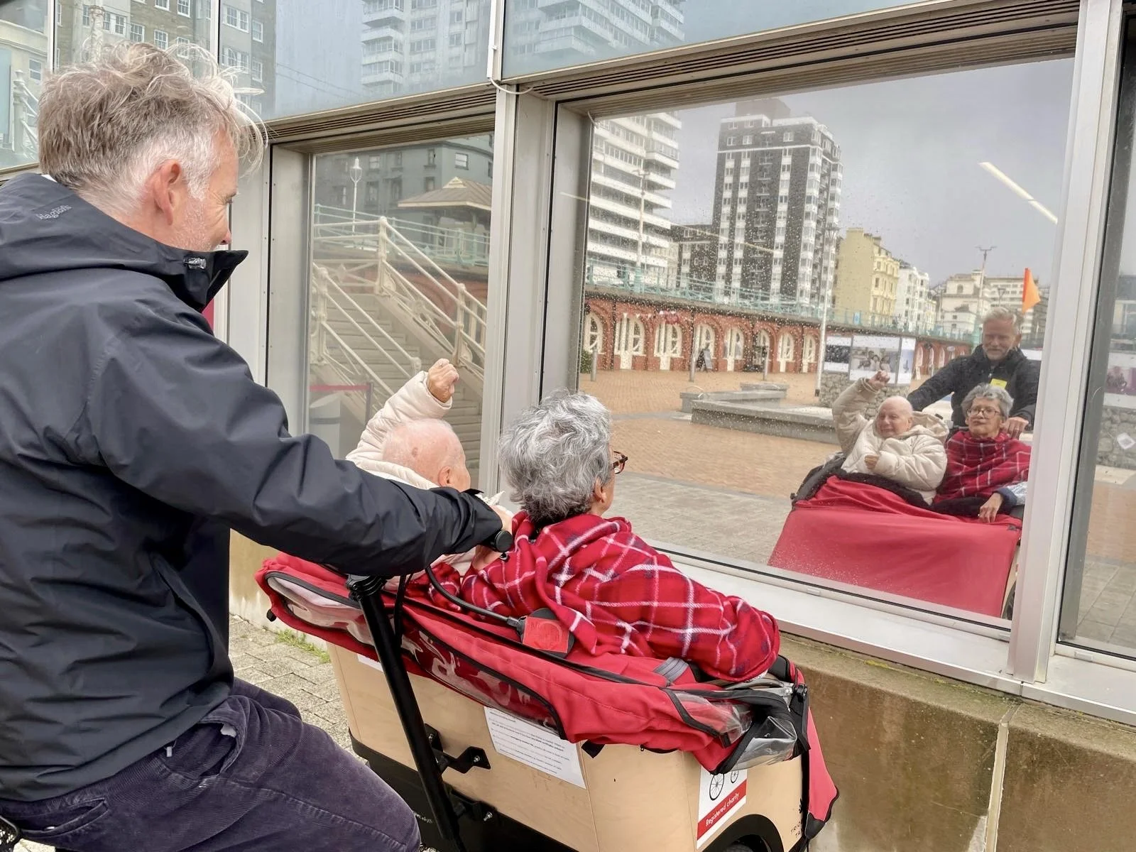 A red trishaw with two participants in the front and the Cycle-Pilot sitting behind. They are looking at their reflection in a window whilst smiling and waving. 
