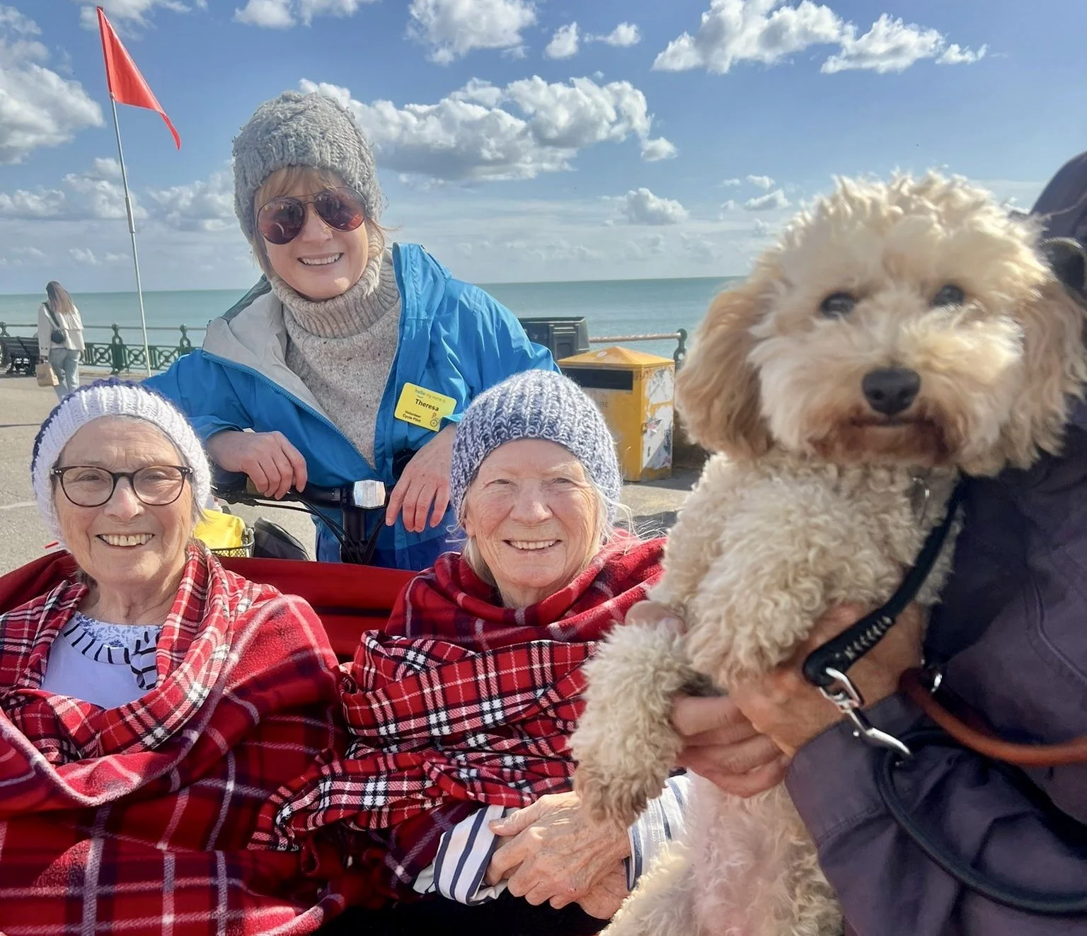 Two participants sit in the front of a trishaw, they are wrapped up in red checked blankets. The Cycle-Pilot sits behind them. In the foreground is a small dog with cream, curly fur being held in someone's arms. Everyone is smiling and looking a the 