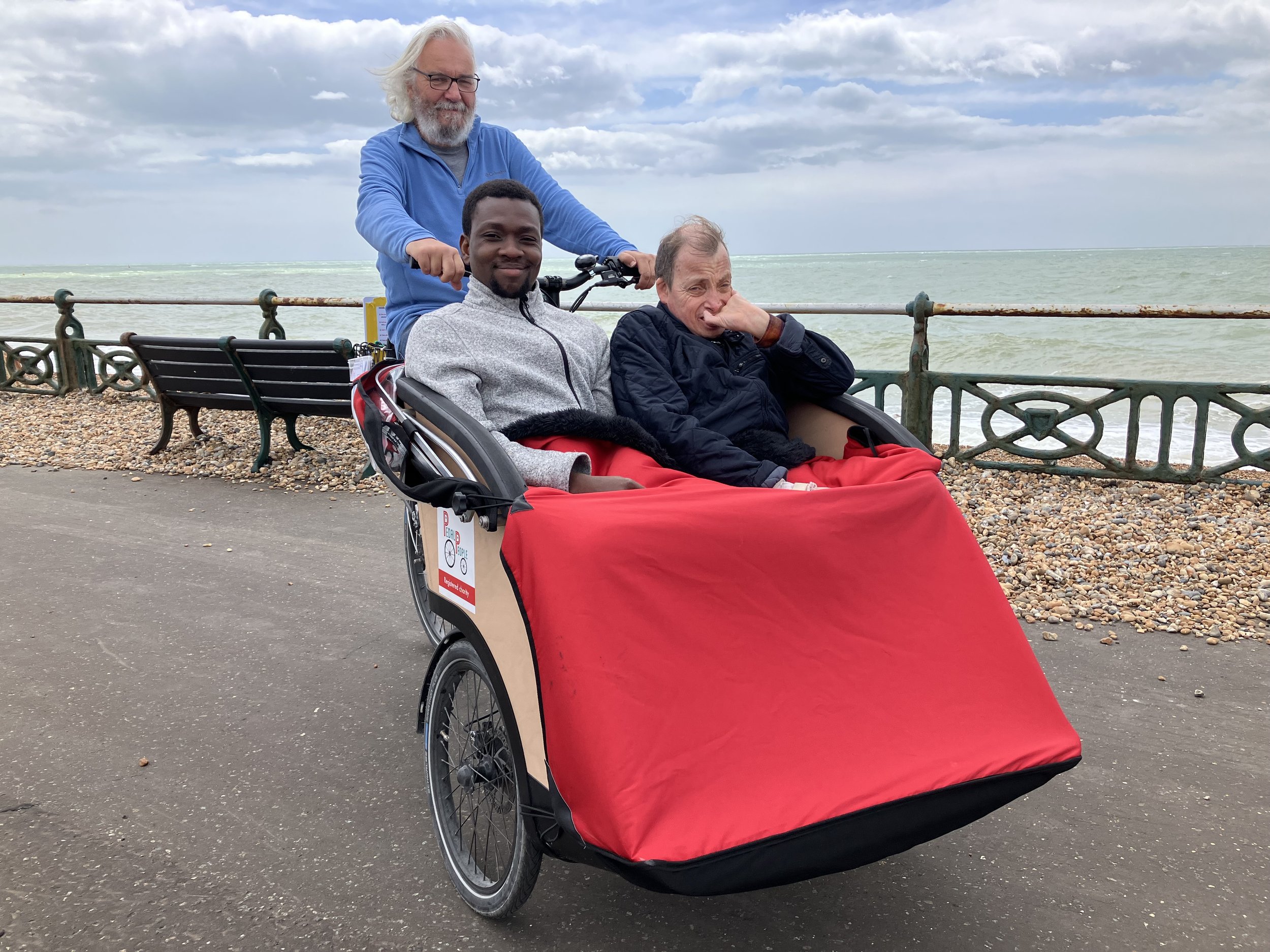 Bright red trishaw cycle with 2 participants seated in the front. Volunteer cycle pilot seated on the back of the cycle. Seaside in background.