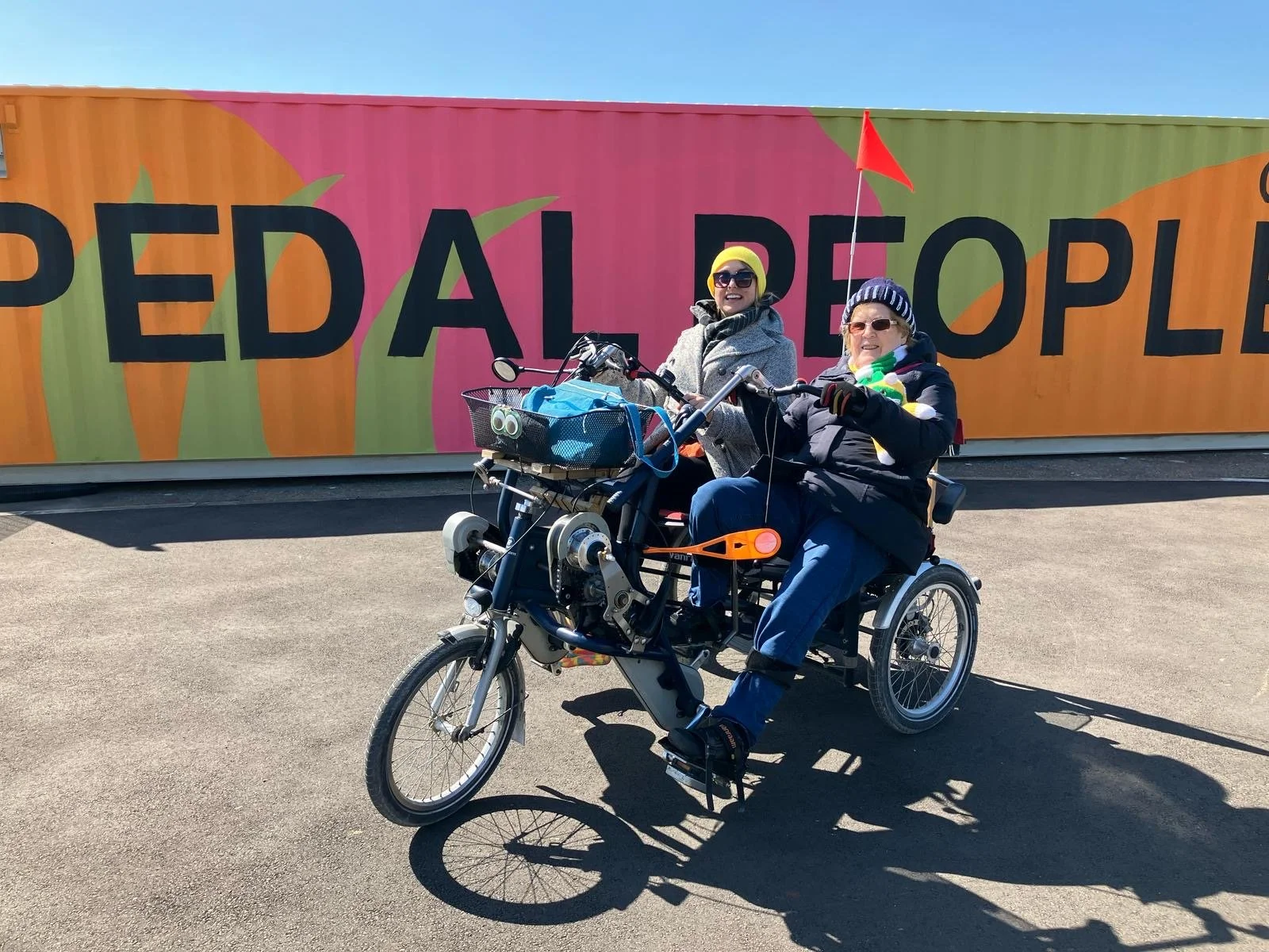 
Side-by-side seated tandem cycle with 2 participants riding. Both smiling at camera. Pedal People base and mural behind them.  