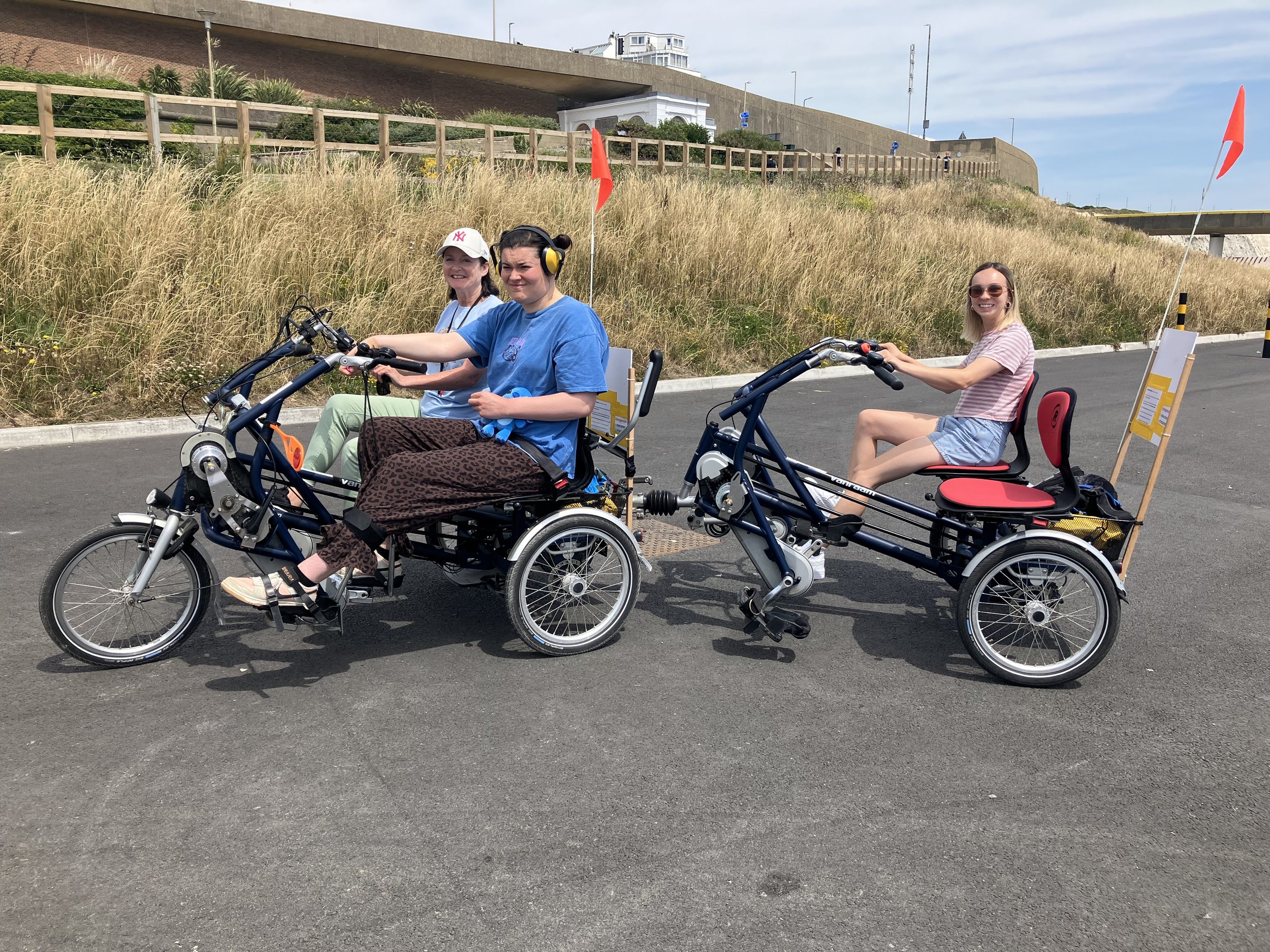 2 side-by-side tandems attached together
to create the 'Fun Train'. 2 Participants sit at the front and one on the back. All smiling. Grassy hill in the background and blue sky.