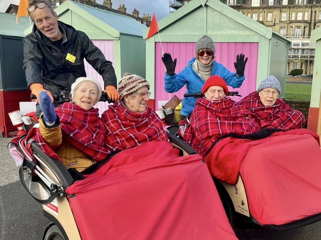Two trishaw bikes parked side by side, each carrying passengers wrapped in red checked blankets. The Cycle-Pilots are sat behind. Colourful beach huts line the background, and historic buildings are visible further back.