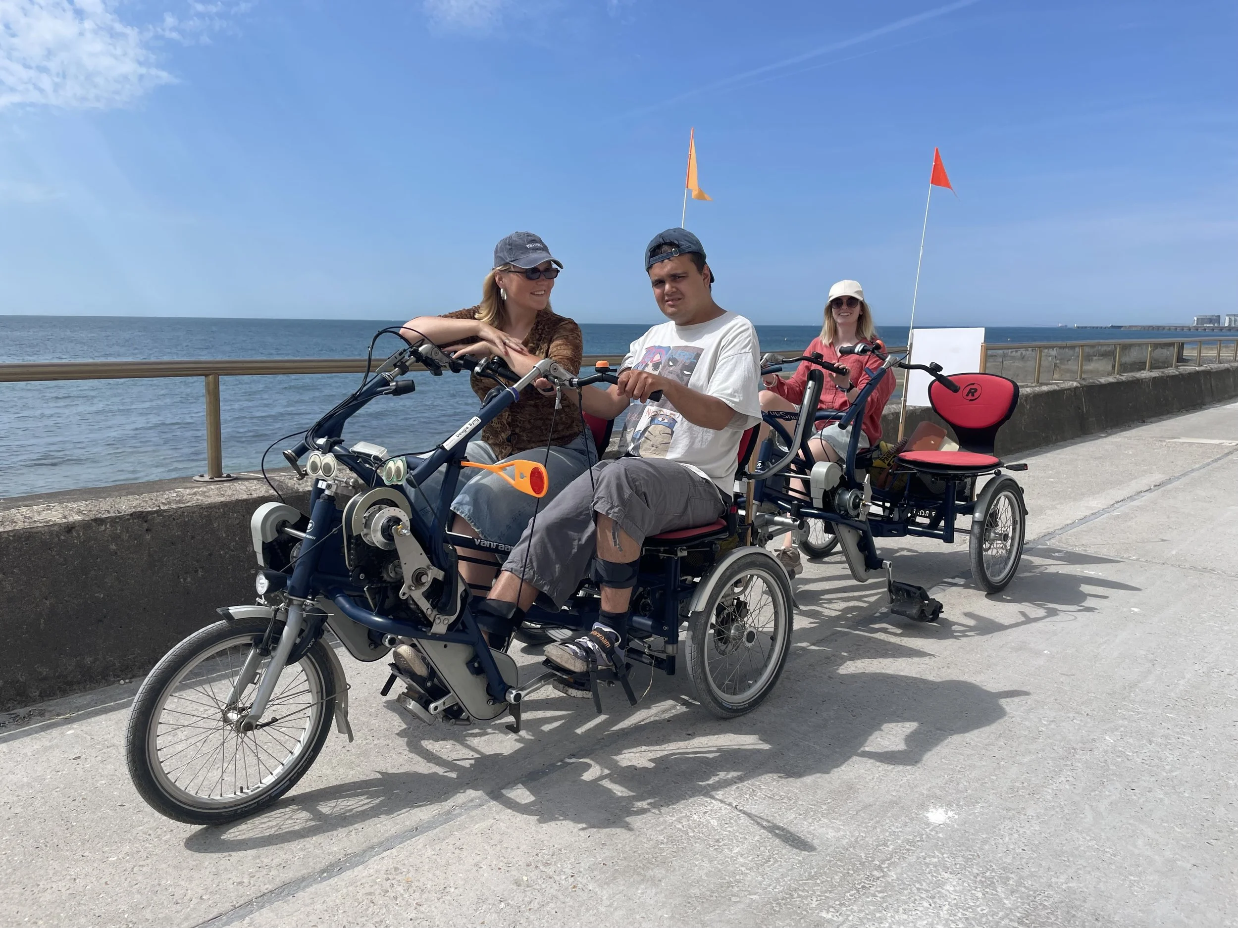 Two people sit on the front of a side-by-side tandem. There is side-by-side tandem tag-along attached behind them with one rider. There is blue sky and sea in the background.