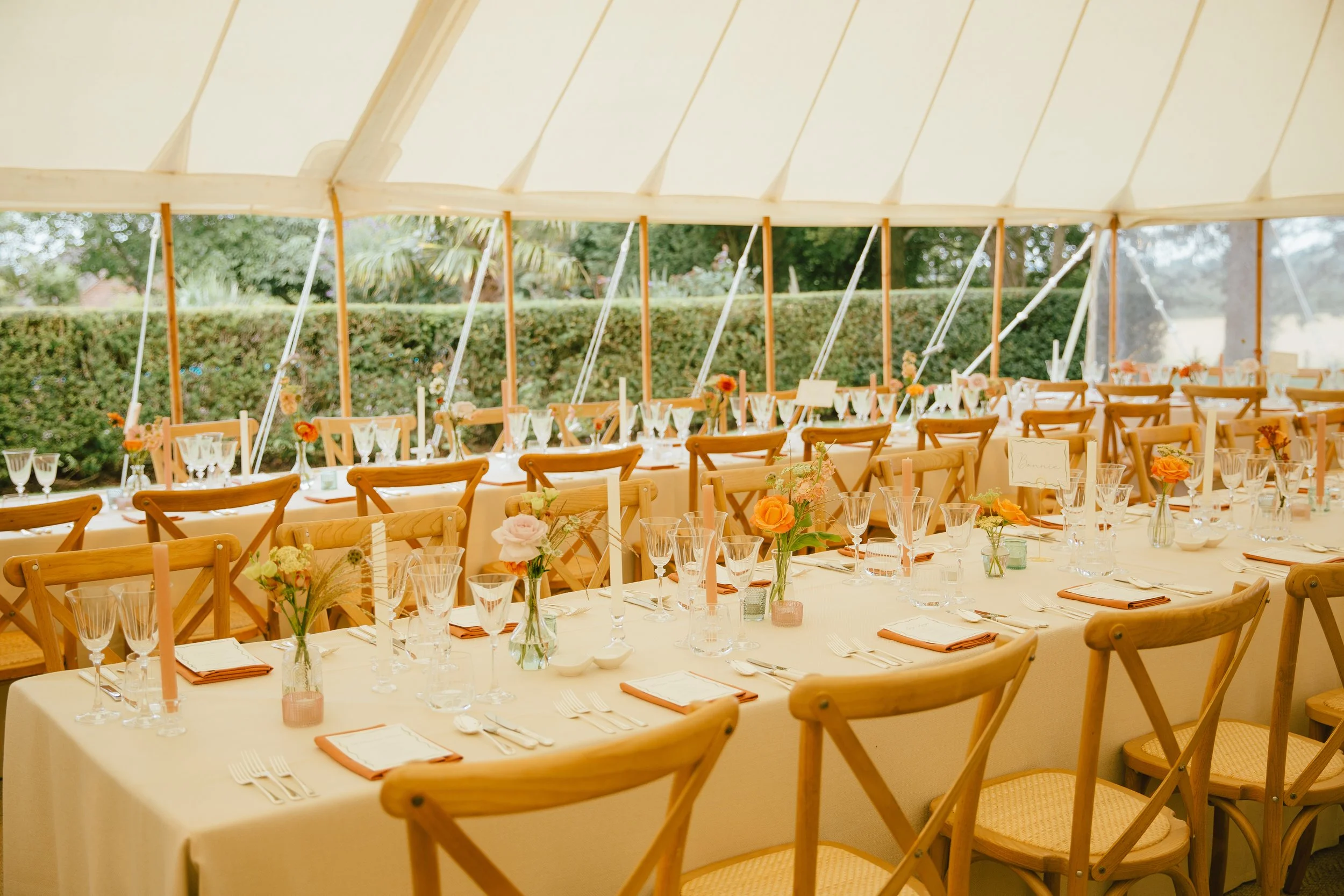 A decorated event tent with long dining tables set with flowers, candles, glassware, plates, and silverware, ready for a celebration.