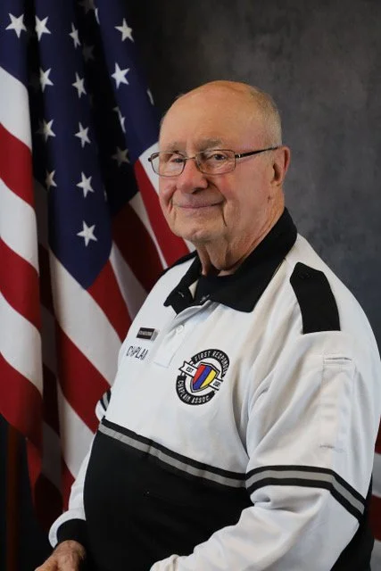 Older man in a white uniform with a black collar and shoulder patches, standing in front of an American flag.