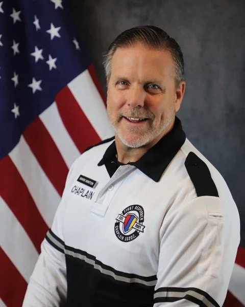 A man in a white and black uniform standing in front of an American flag. He is smiling. The uniform has patches and a name tag that reads 'Corneal'.