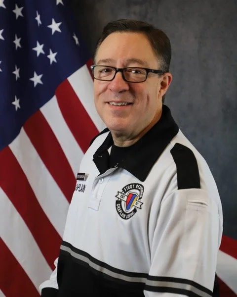 A man with glasses and a beard, wearing a uniform, standing in front of an American flag.