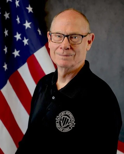 A man with glasses wearing a white and black uniform, standing in front of an American flag.