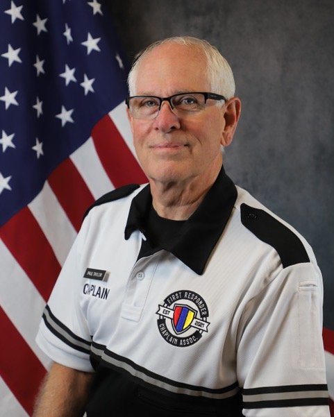 A man in a chaplain uniform smiling, with an American flag in the background.