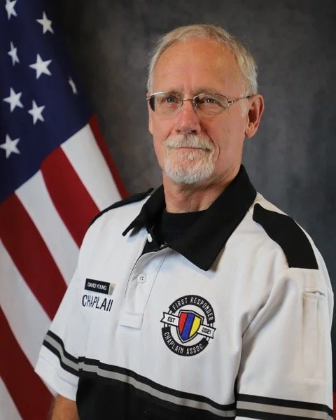 A man with gray hair and a beard wearing a white and black sports jacket, standing in front of an American flag.