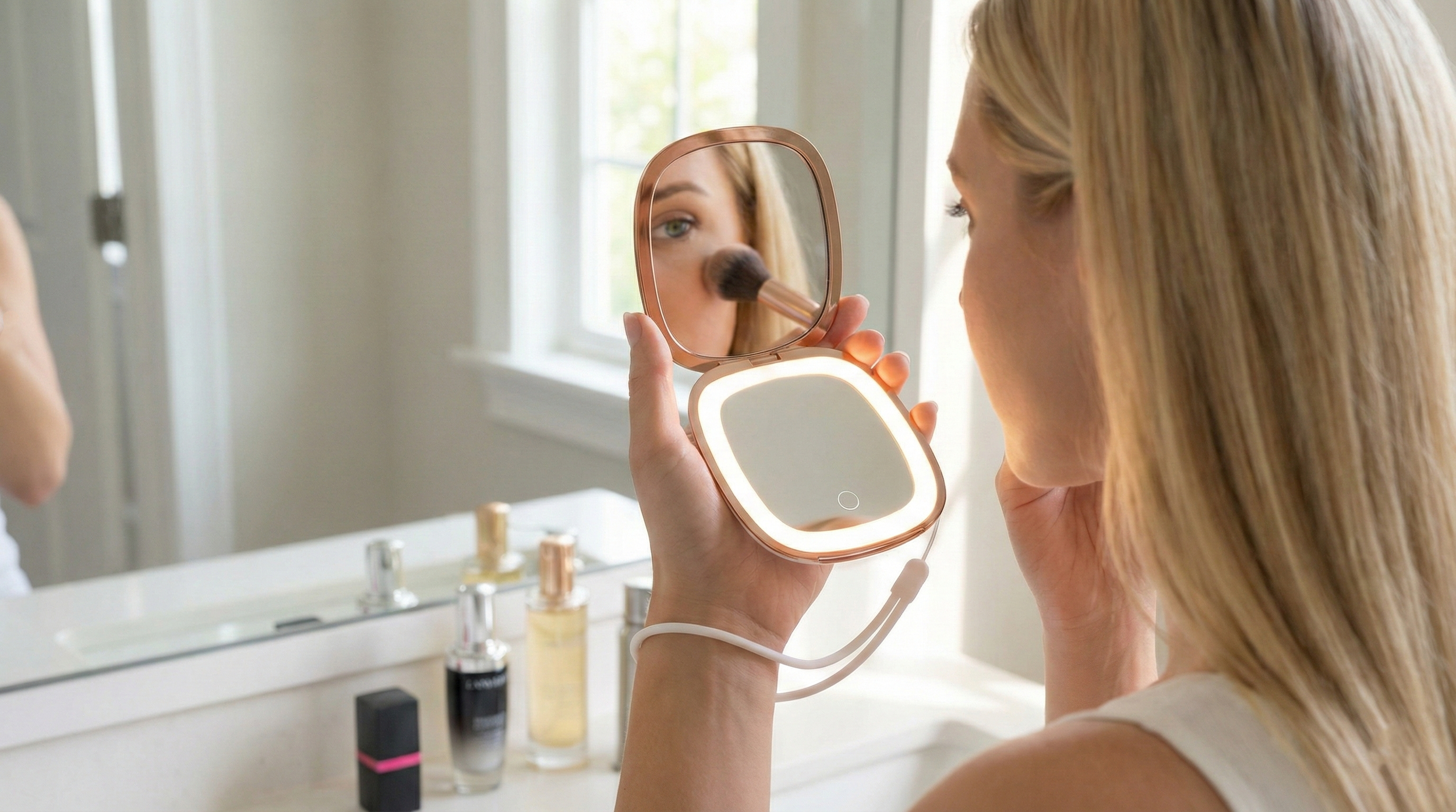 A woman applying makeup, looking into a small illuminated mirror, with a brush in her hand and skincare bottles on the bathroom counter.