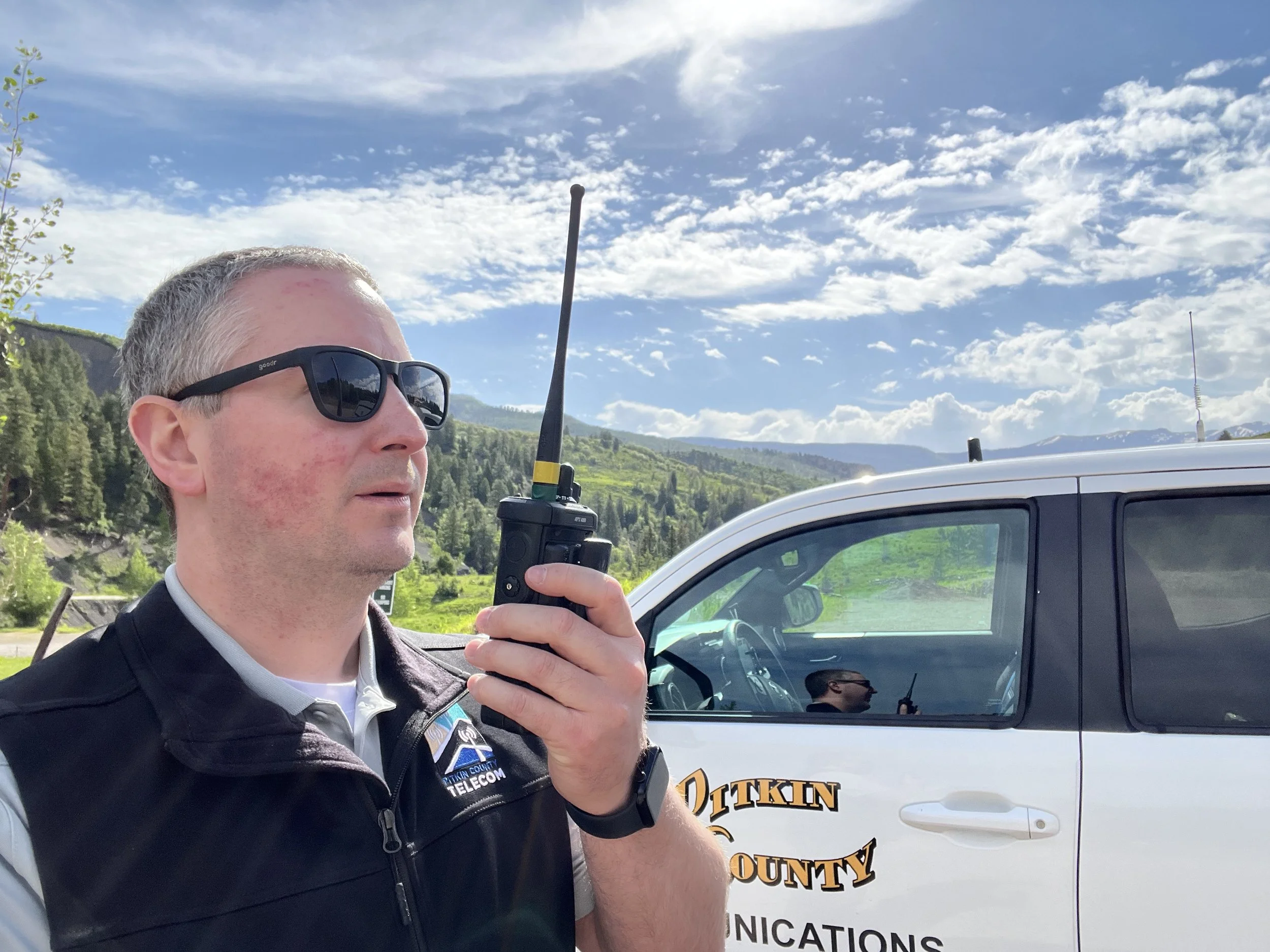 man talking into a handheld radio with a logoed truck and mountains in the background