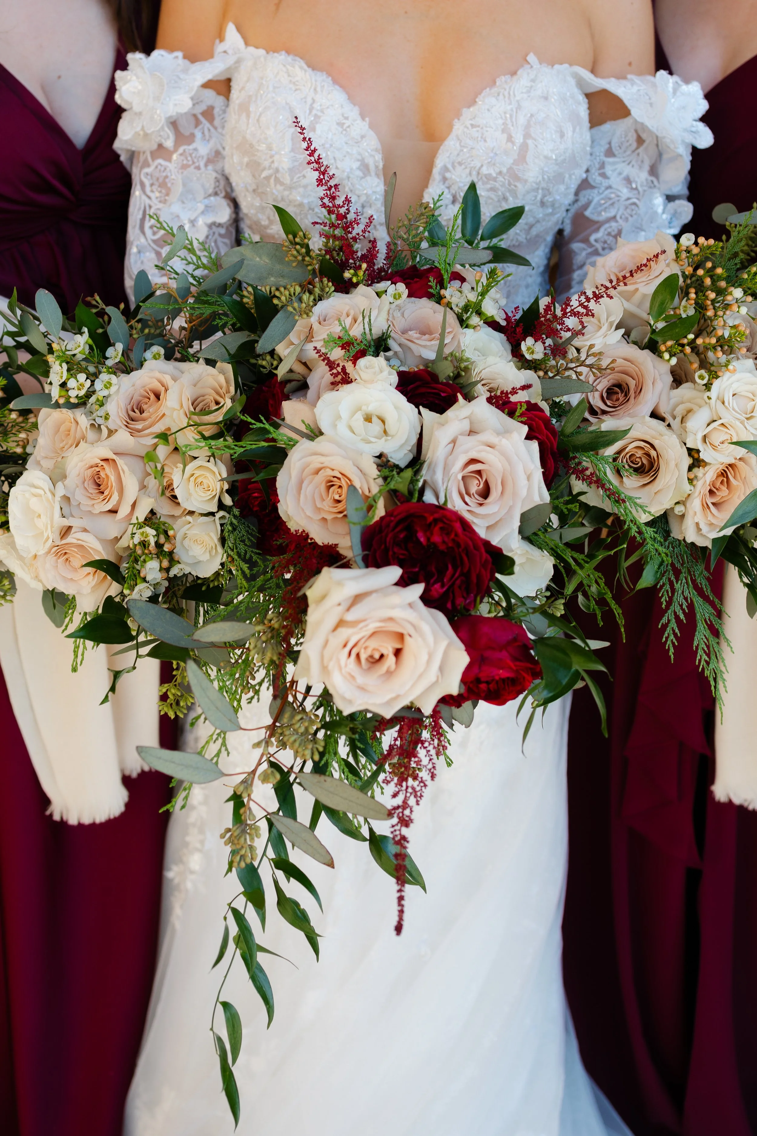 Bride holding a cascading bouquet of cream, blush, and deep red roses with greenery, wearing a lace wedding dress with off-the-shoulder sleeves, flanked by bridesmaids in burgundy dresses.