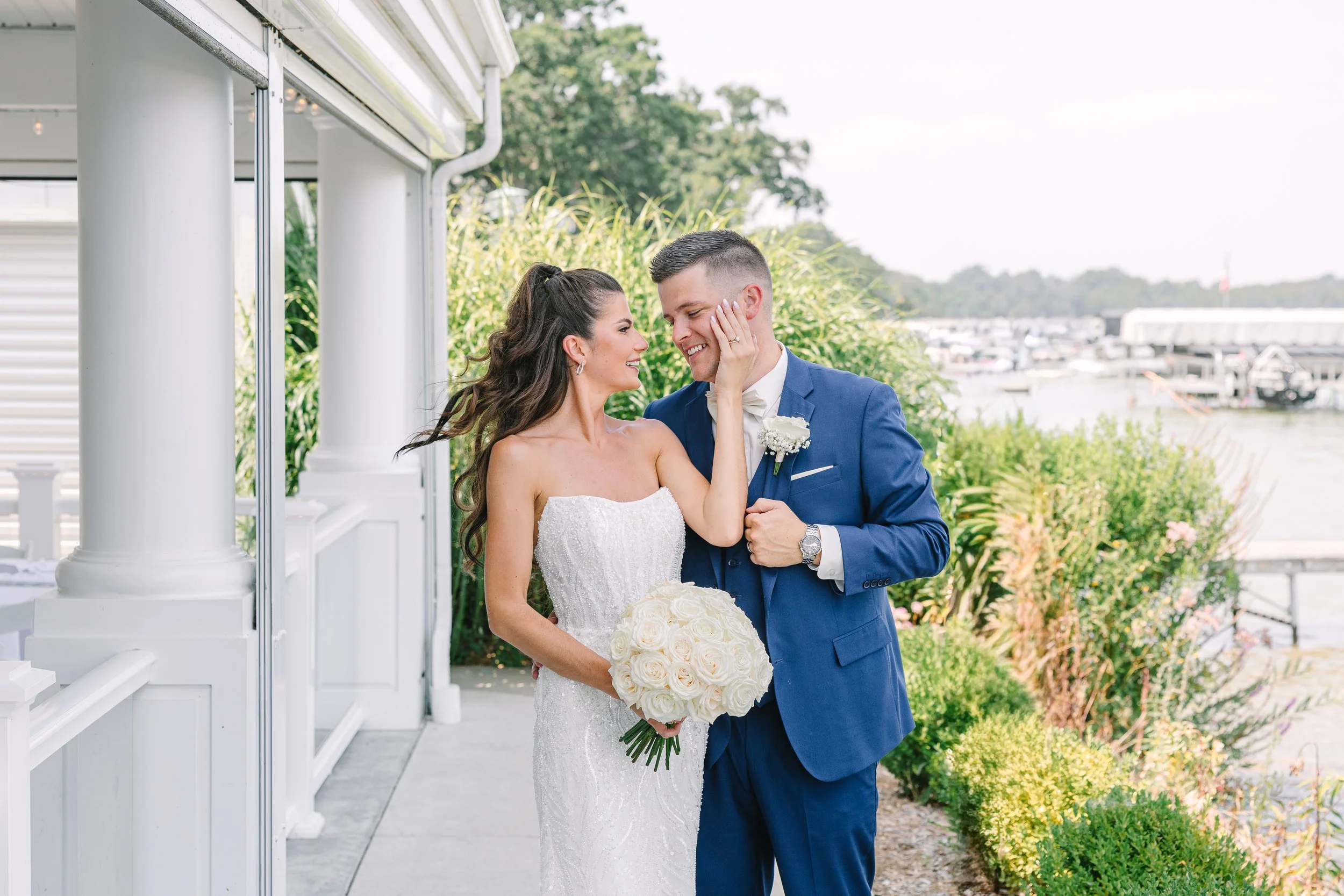A newlywed couple smiling and gazing into each other's eyes on a waterfront outdoor wedding venue with greenery and boats in the background. The bride is holding a bouquet of white roses, and the groom is touching her face.