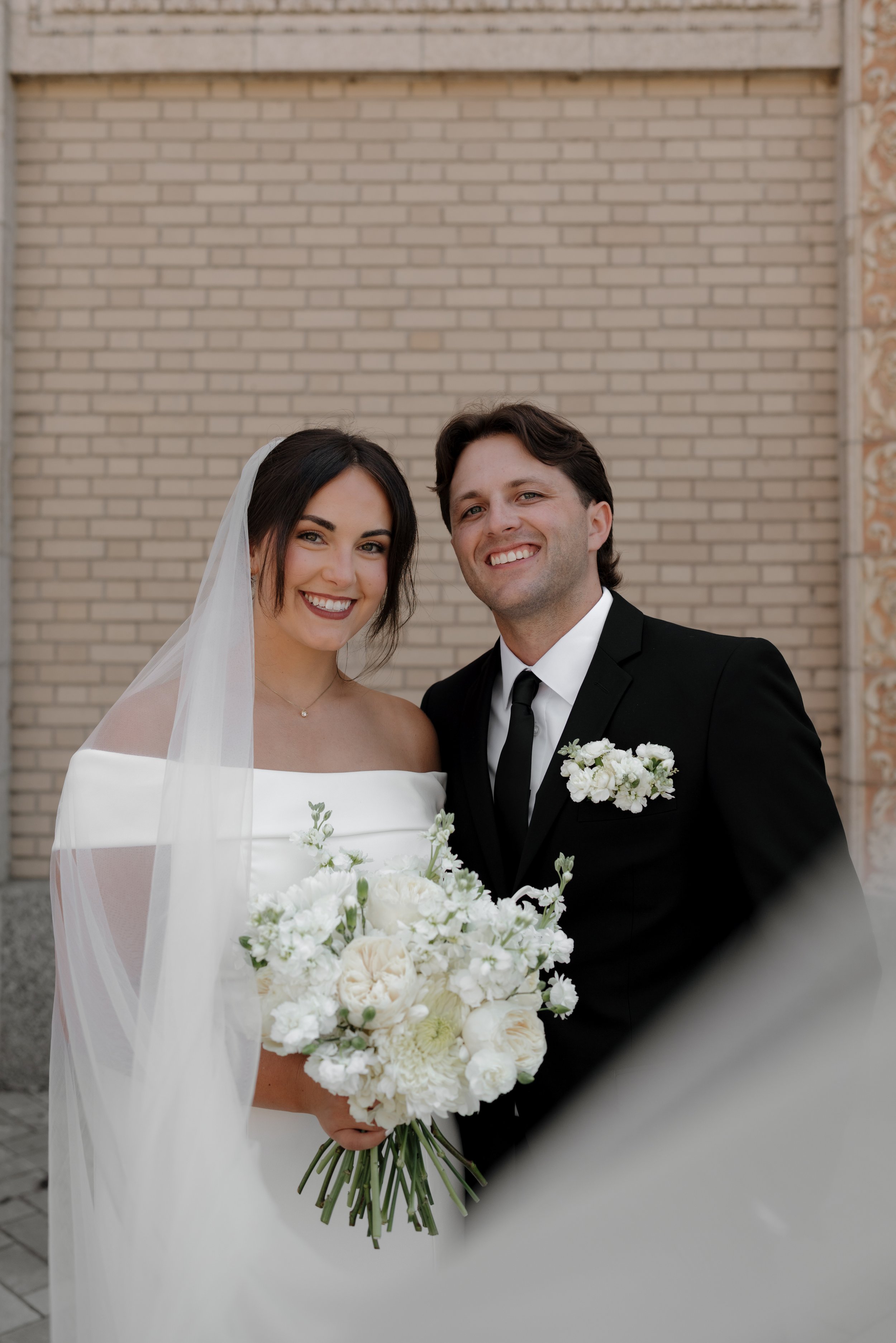 Bride and groom smiling for a wedding photo, with a brick wall background. The bride is in a white off-shoulder wedding gown and veil, holding a bouquet of white flowers. The groom is in a black suit with a white shirt and black tie, with a small whi
