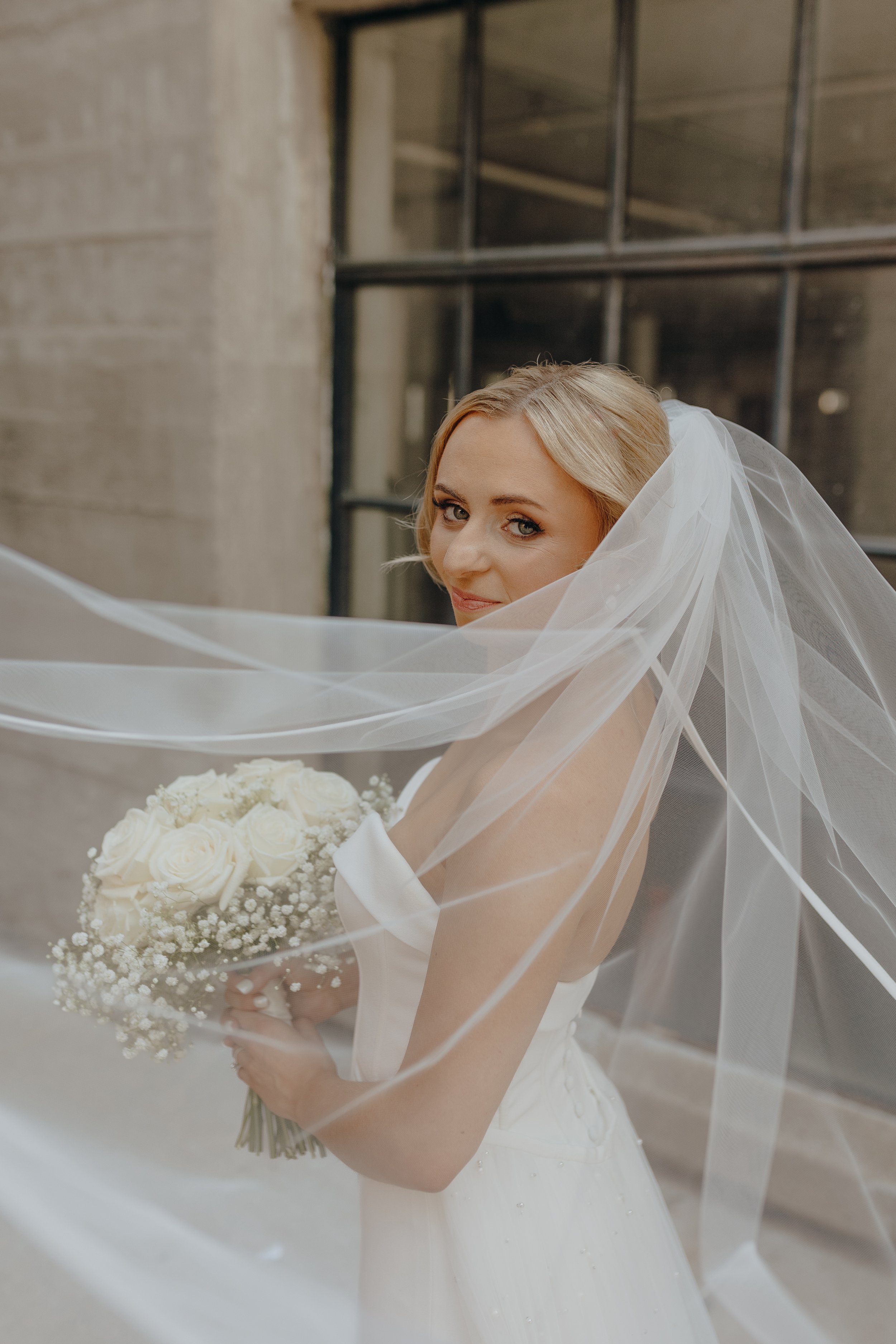 Bride in a wedding dress holding a bouquet of white roses, with a sheer veil covering part of her face, standing in front of a window.