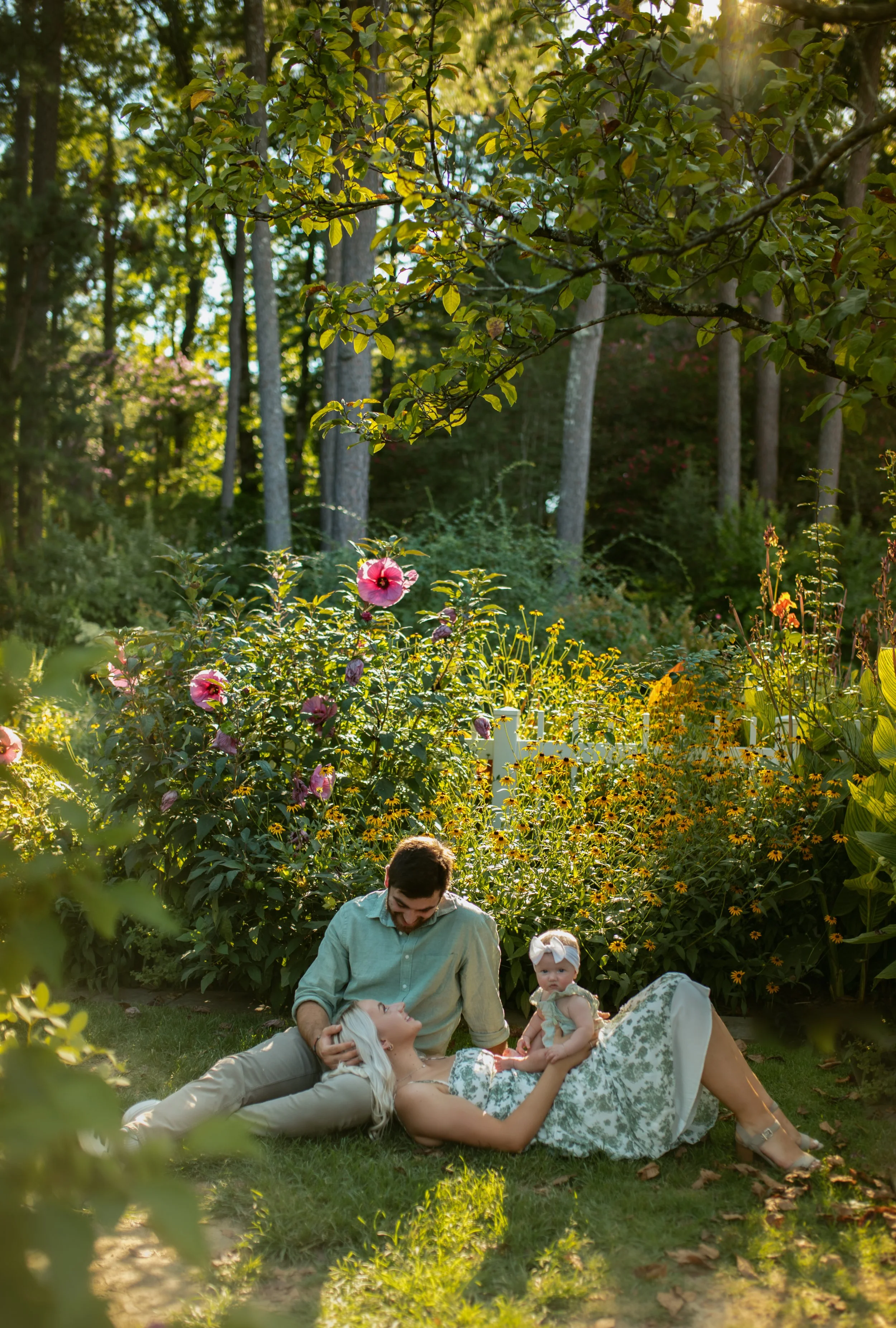 A family of three in a lush garden with colorful flowers, including pink and yellow blooms. The man is sitting on the grass, smiling and leaning towards the woman who is lying down with her head on his lap. The woman has long blonde hair and is dressed in a floral dress. Their young daughter, wearing a light green dress and a white headband, sits beside her mother.