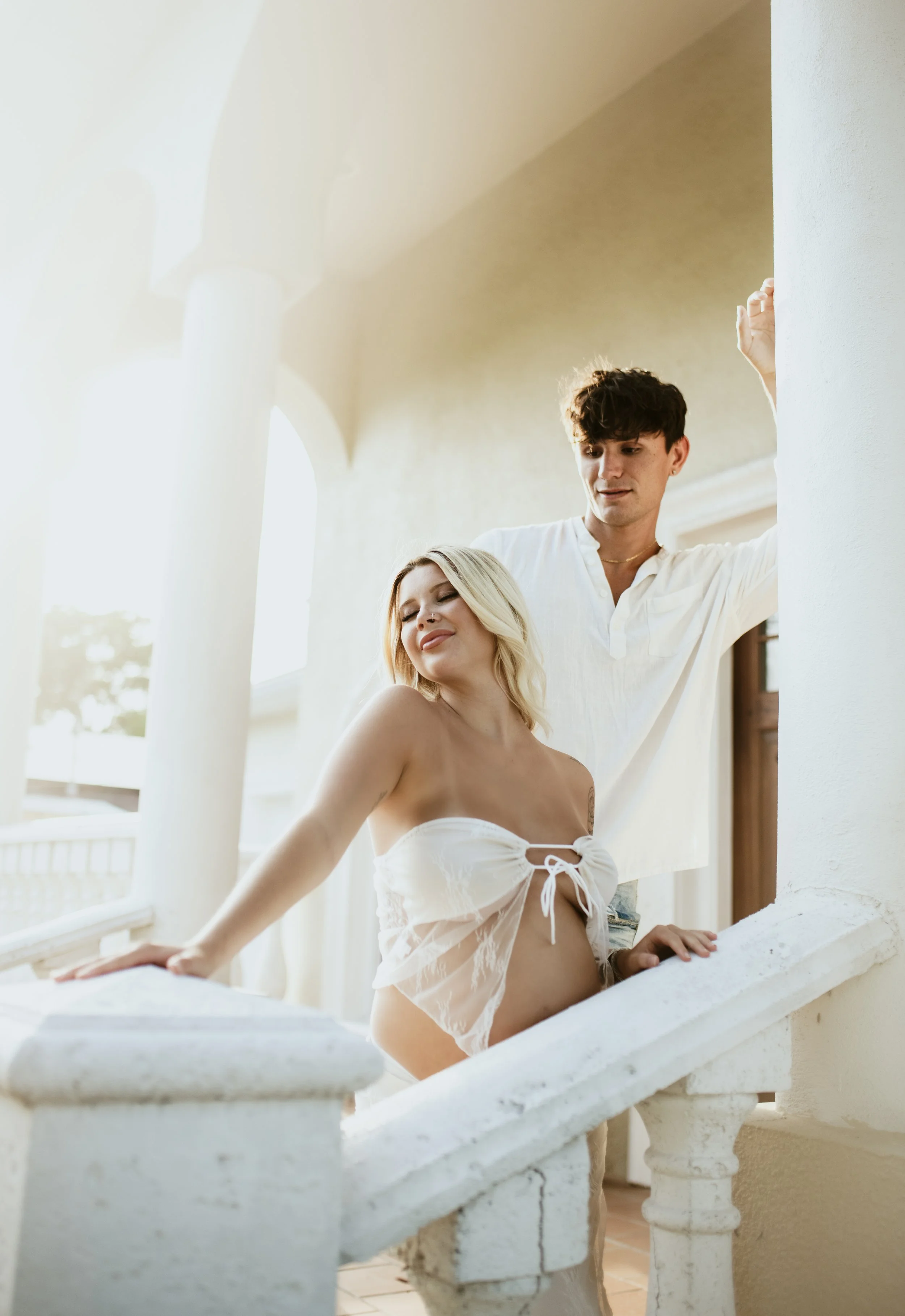 A young couple stands on a white balcony, with sunlight streaming in, intimate and content.