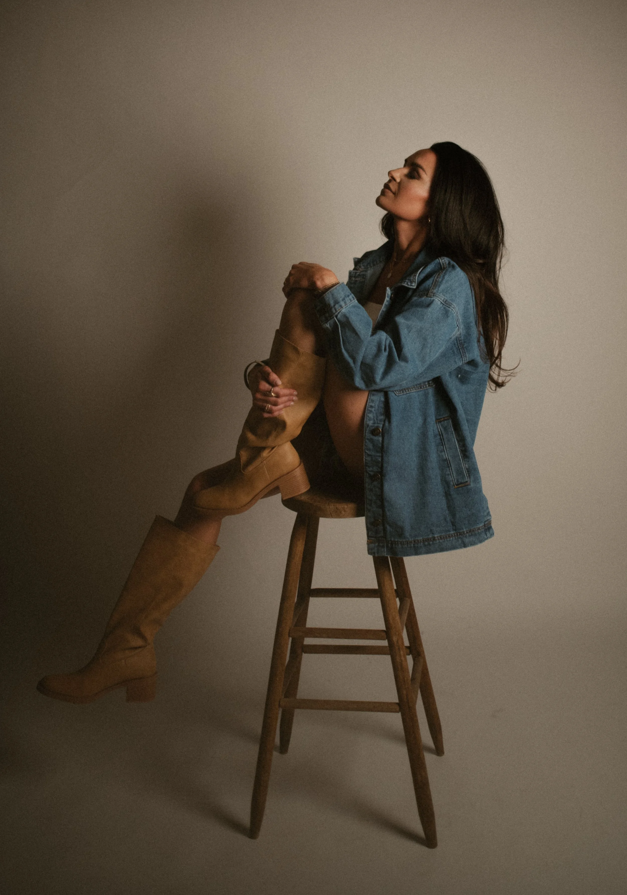 A woman with long dark hair sits on a wooden stool, wearing a denim jacket, black underwear, and tan knee-high boots, against a neutral background.