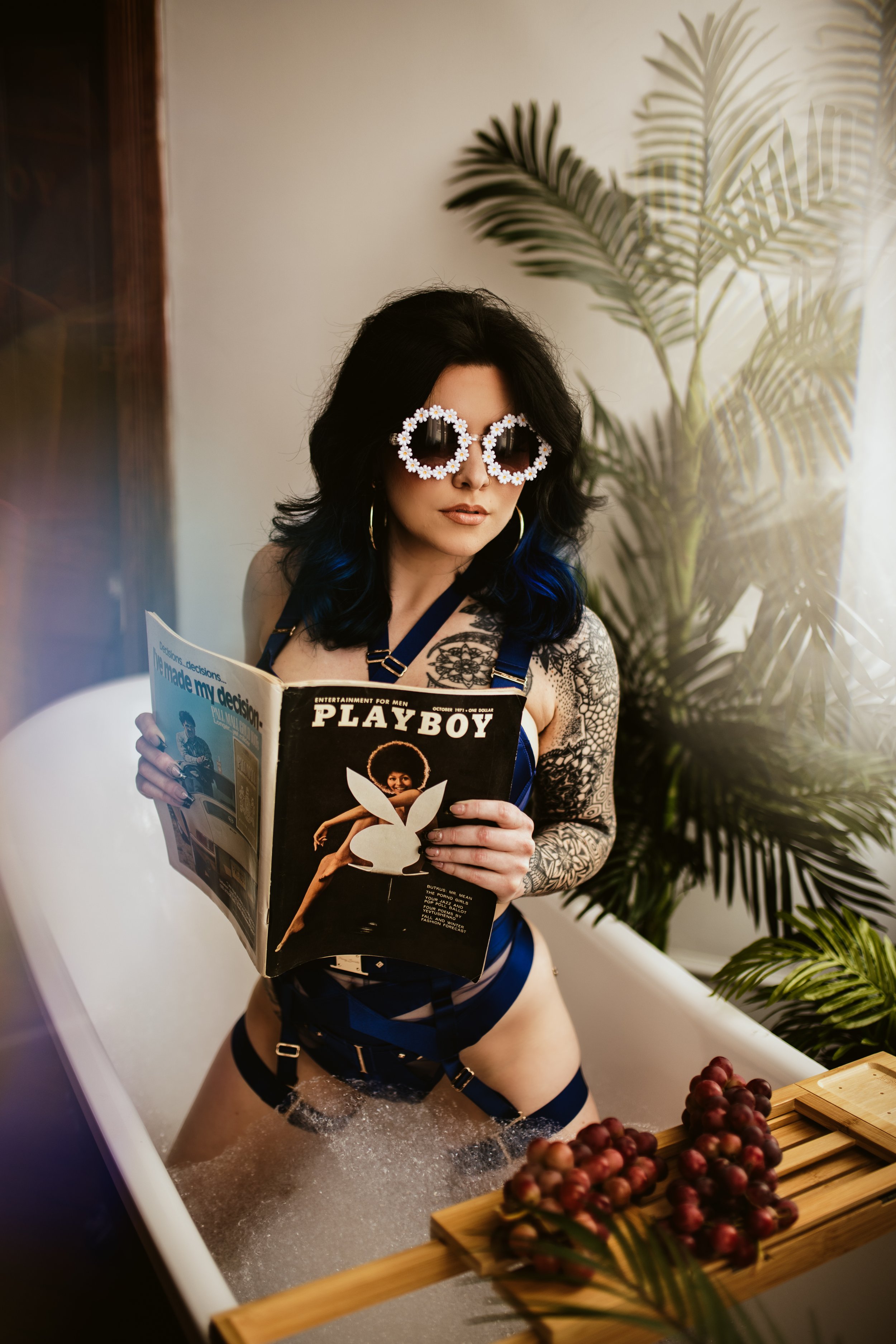 Woman with black hair, sunglasses decorated with flowers, and tattoos, sitting in a bathtub with water and bubbles, holding a Playboy magazine, with grape clusters on a wooden tray in front of her and tropical plants in the background.