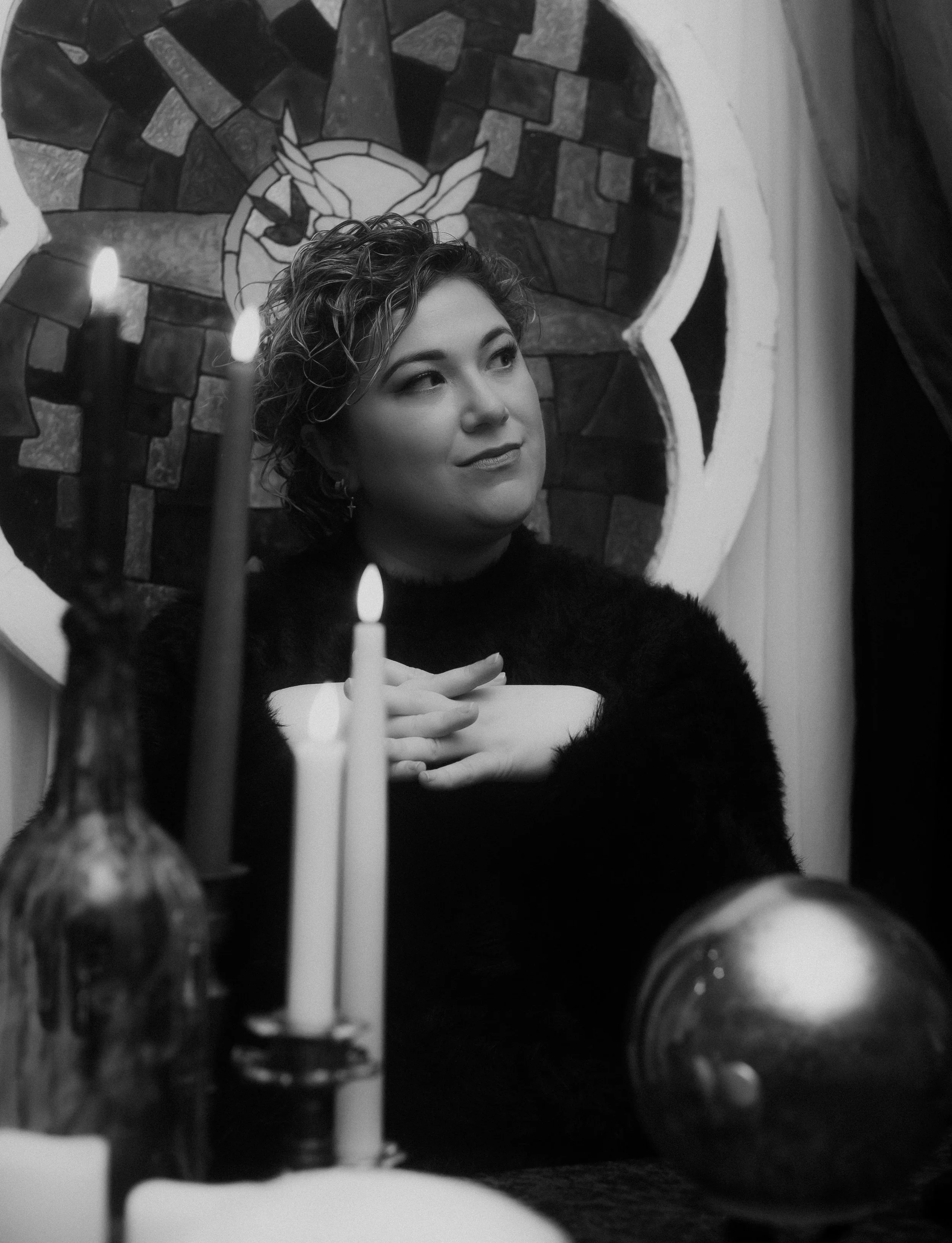 Black and white photo of a person holding a candelabra, surrounded by lit candles. They wear a spiked crown and raise one hand upwards. A stained-glass window with a dove is in the background. Items like a crystal ball and skull are on the table.