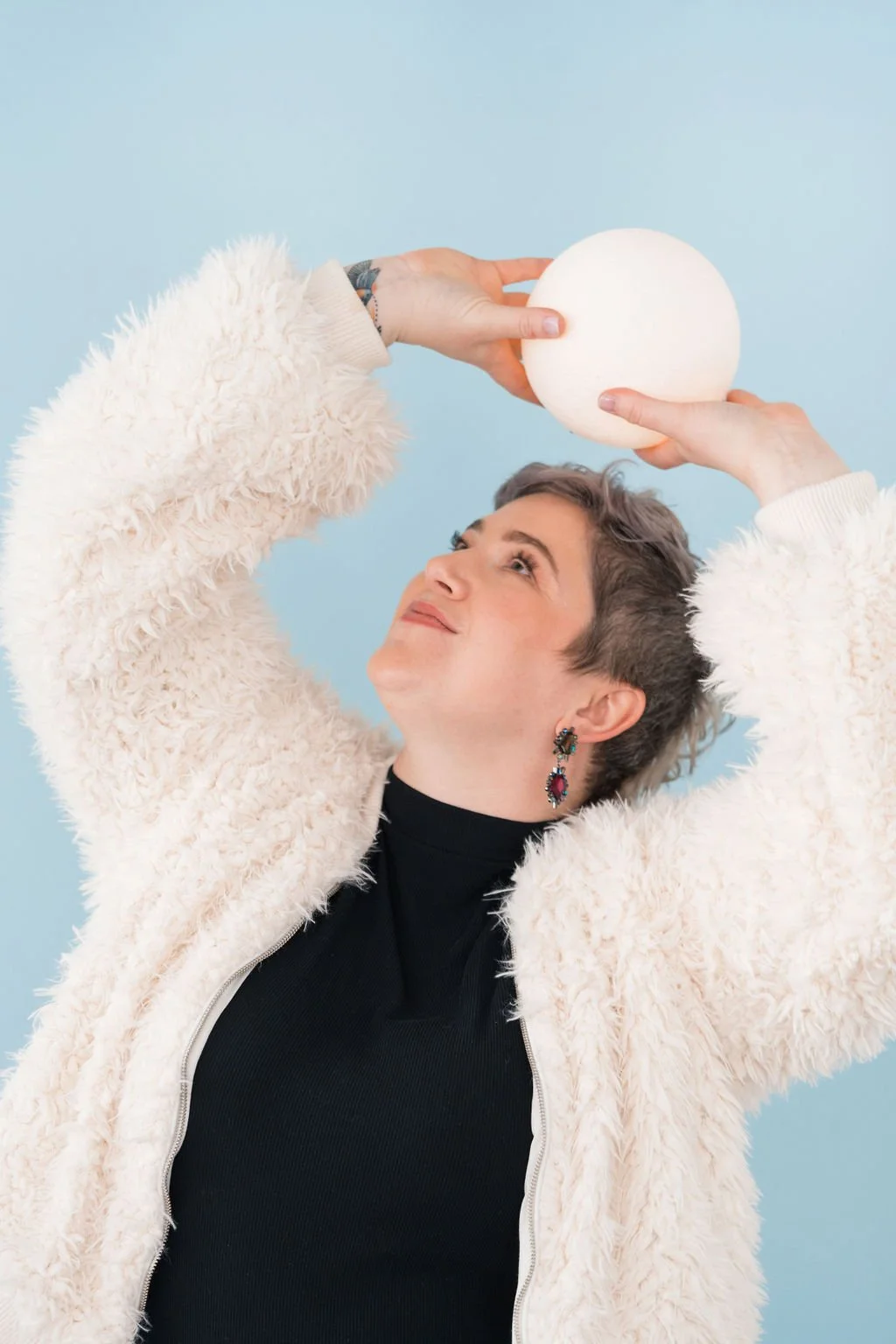 Person wearing a fluffy white jacket holds a white sphere above their head against a blue background.