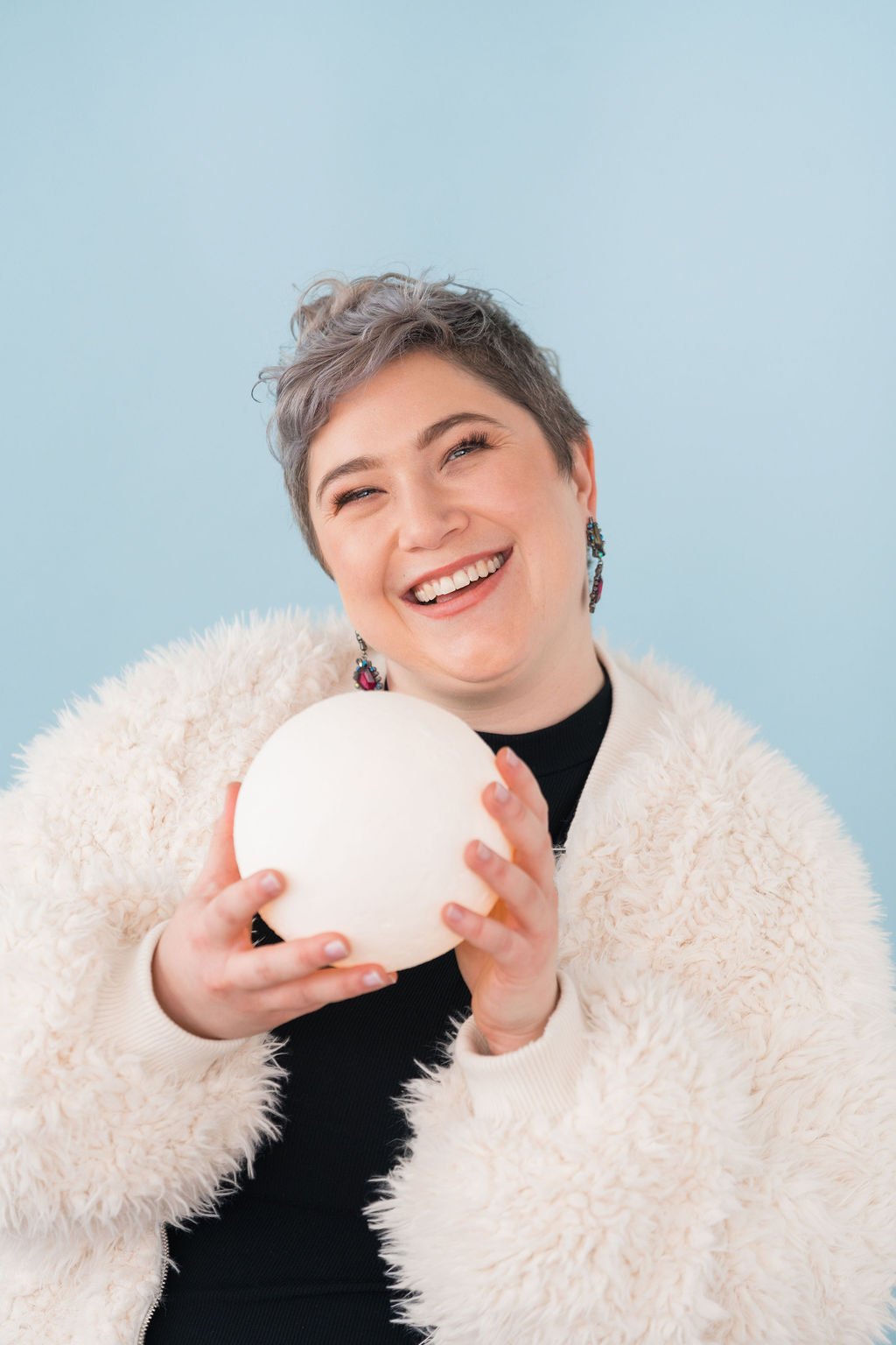 A person with short hair smiling and holding a white ball, wearing a fluffy white jacket and black top against a light blue background.
