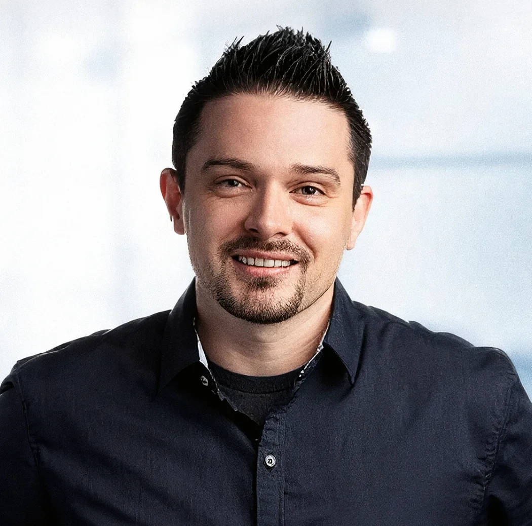 Professional headshot of a smiling man in a dark shirt against a soft light-blue blurred background
