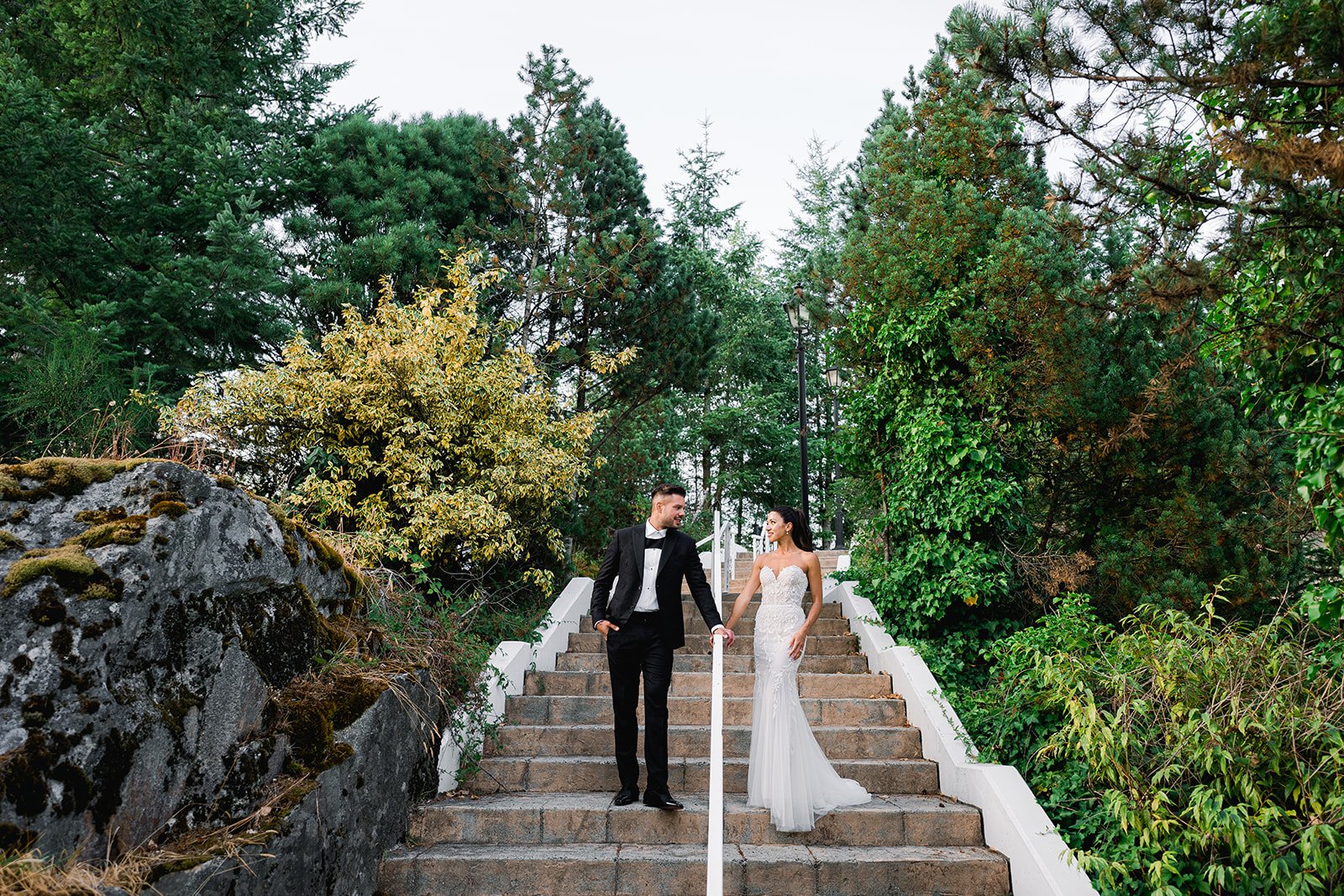 A bride and groom pose on an outdoor staircase surrounded by greenery.