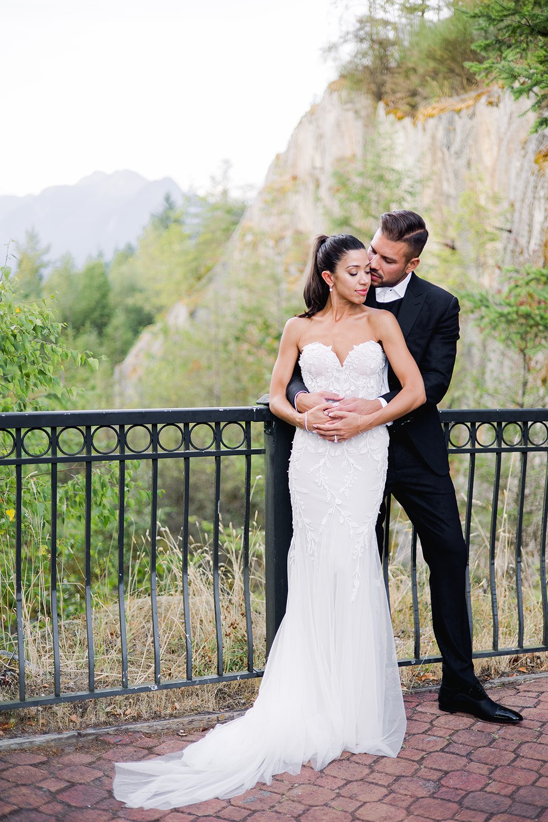 A groom hugs his bride from behind as BC mountains loom in the distance.