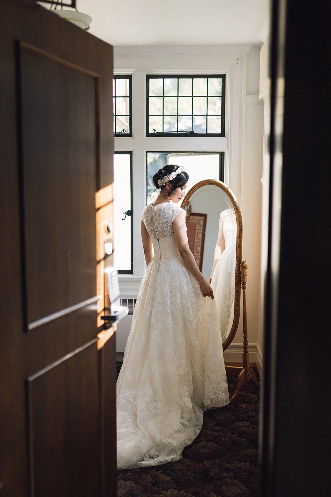 Asian bride in her lace wedding gown looking in a vintage mirror on the morning of her wedding at Cecil Green Park House. Beautiful Life Studios BC.