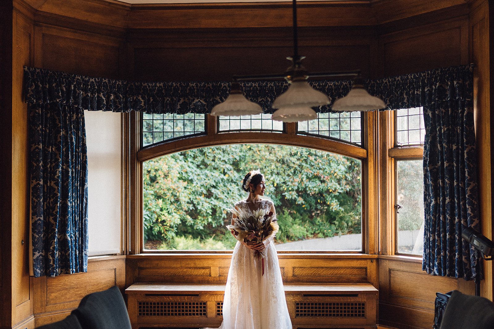 asian bride holding her fall-inspired bouquet in front of a big window at Cecil Green Park House in Vancouver, BC.