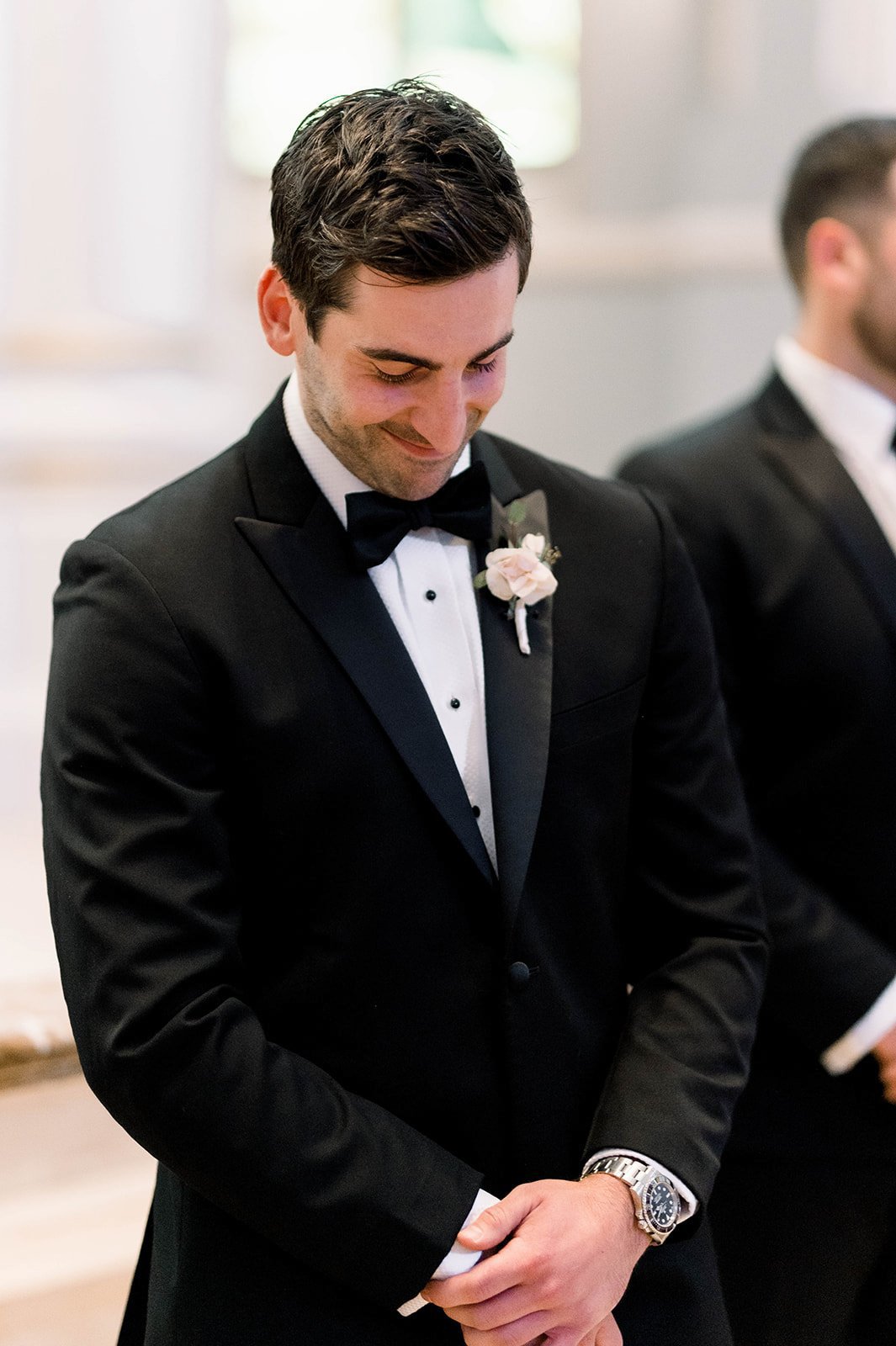 Groom smiles with hands clasped, his eyes cast downward.