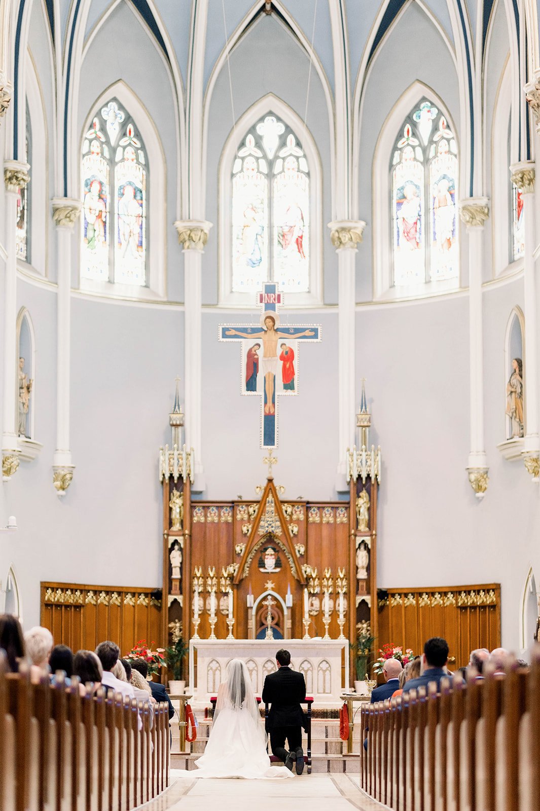 Bride and groom knee at front of aisle in front of the priest and large cathedral windows in hart house ceremony 