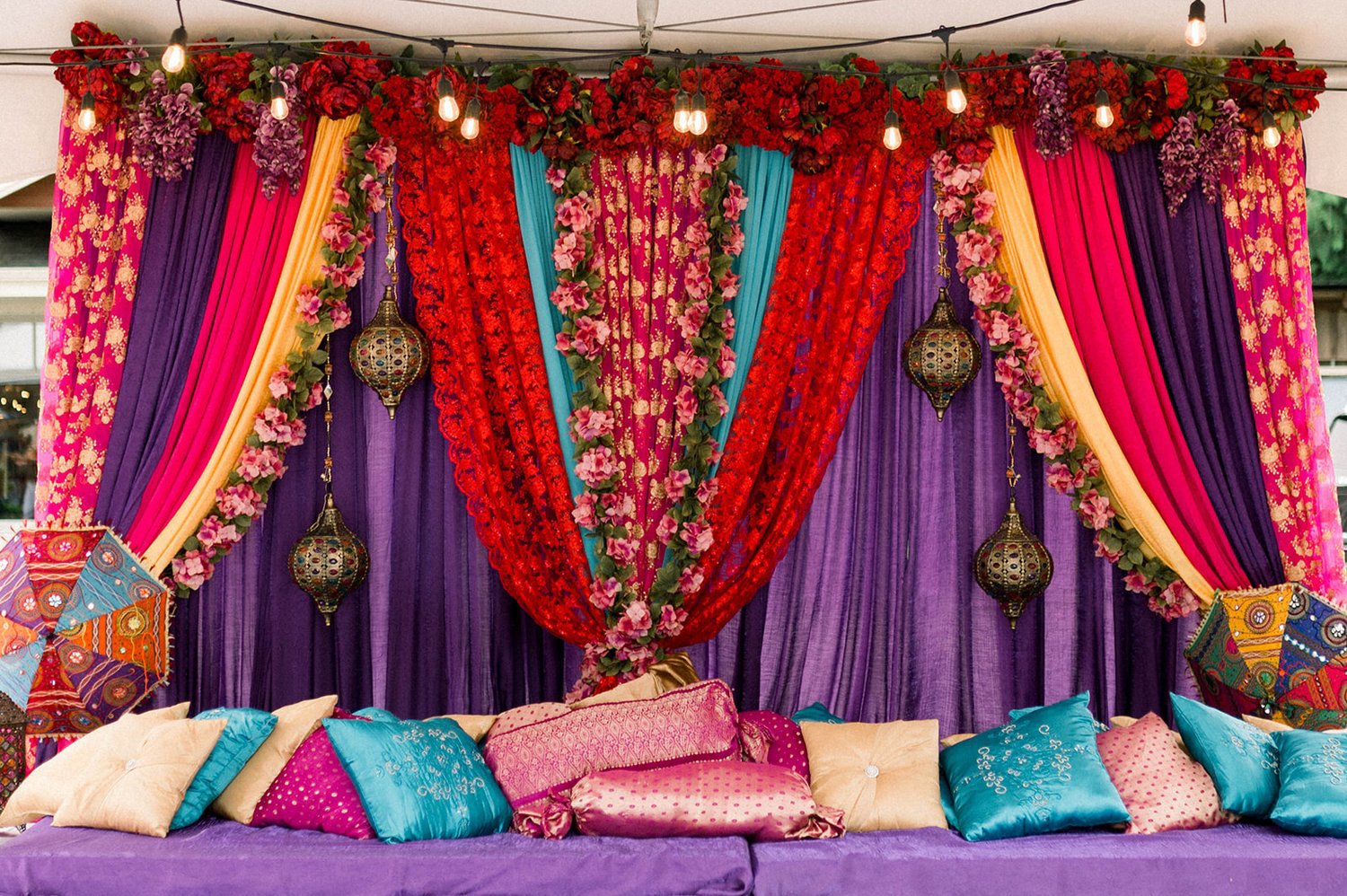 Jewel encrusted lanterns hand between colorful curtains and flowers behind a purple stage covered in silky bright pillows at Sikh Wedding.