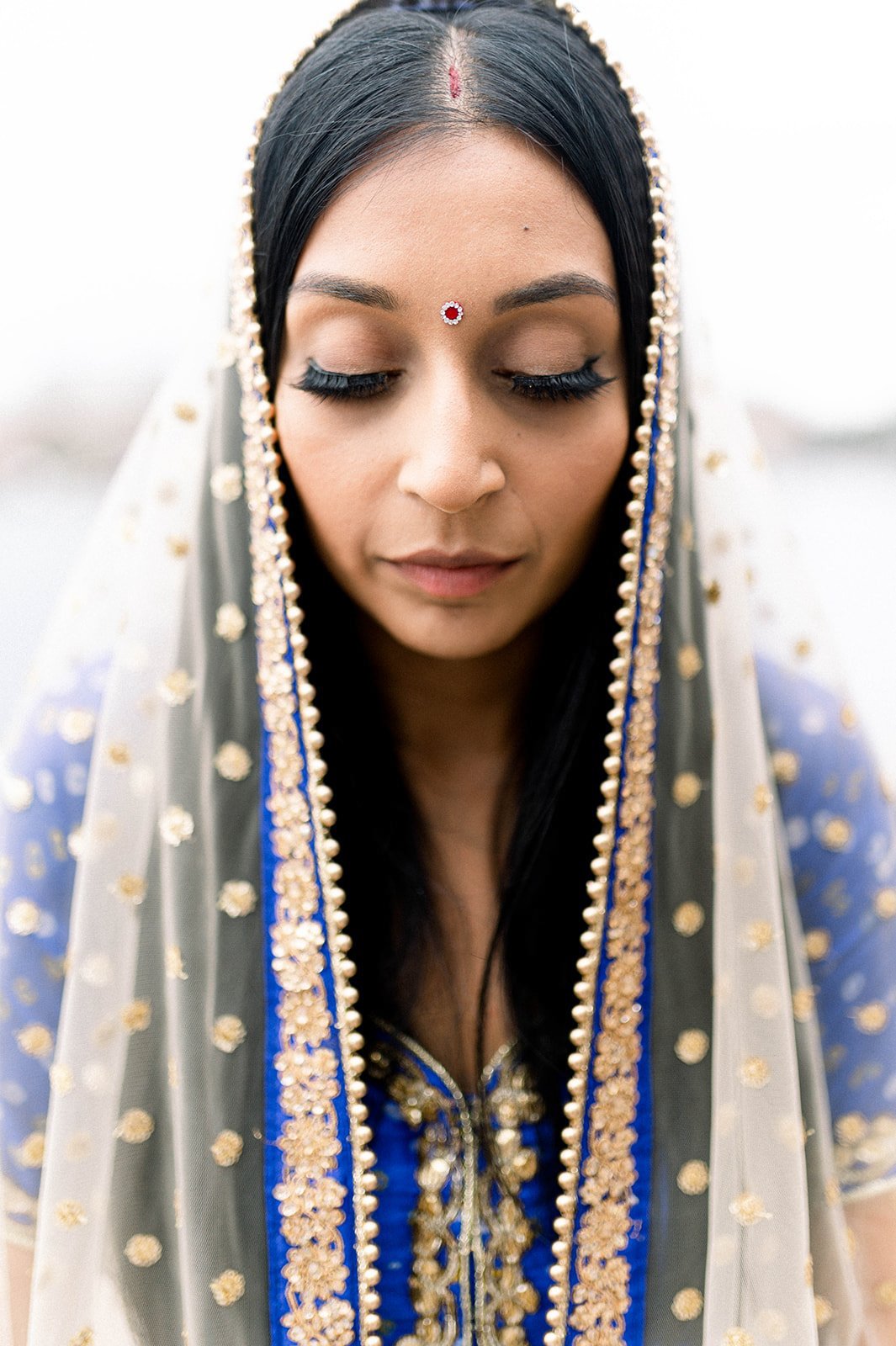 An Indian Bride dressed in blue sits with her eyes closed at her South Asian Wedding. 