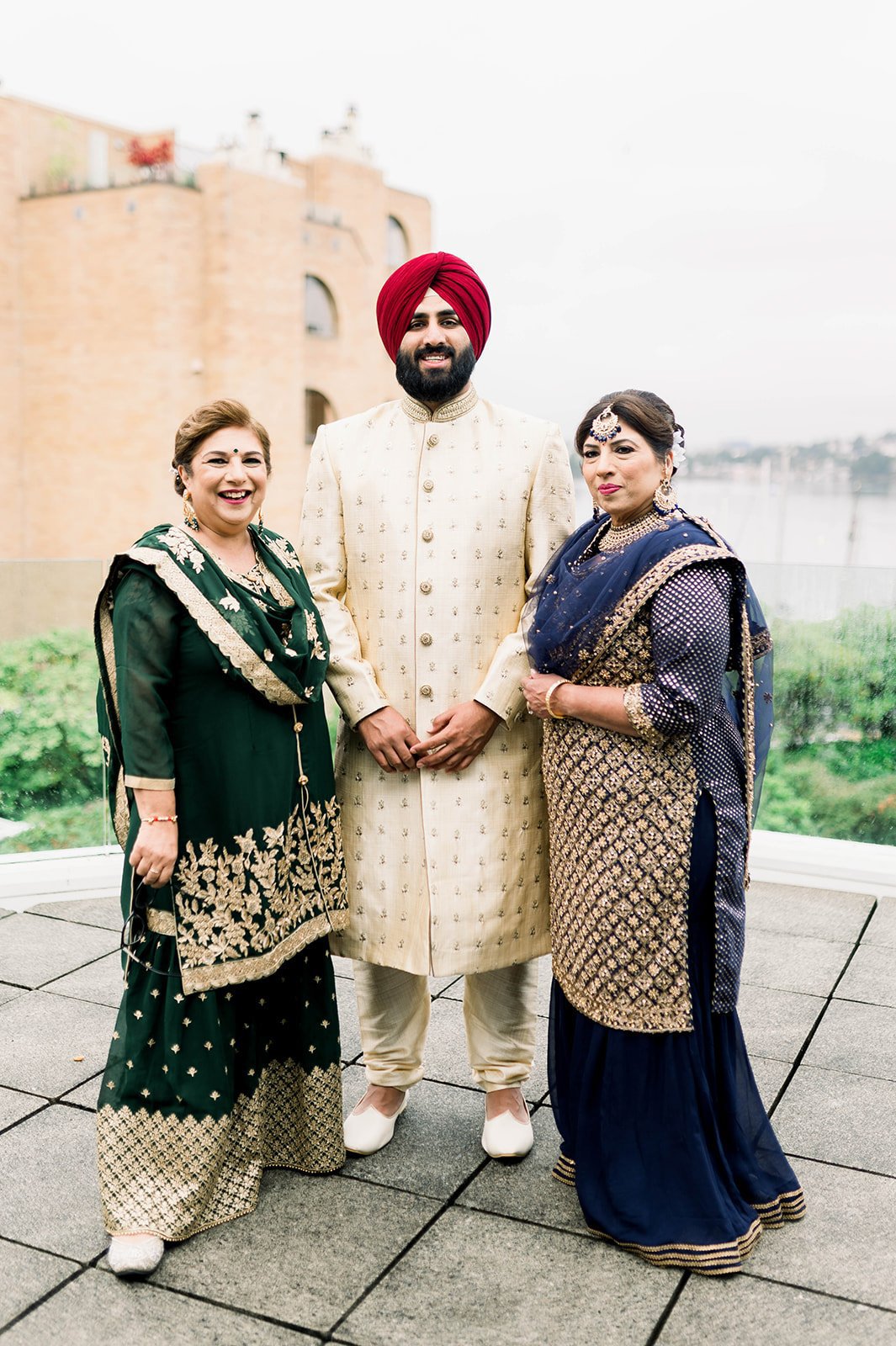An indian groom in a red turban and cream coat smiles with his Aunts before getting married in Victoria BC.