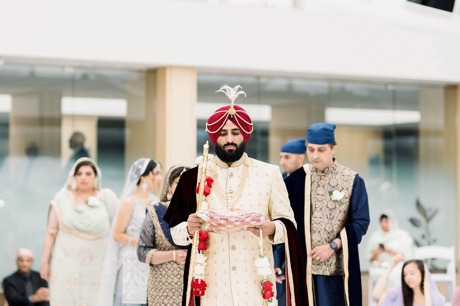 A South asian groom in a red turban and cream coloured coat is escorted in the temple for his wedding