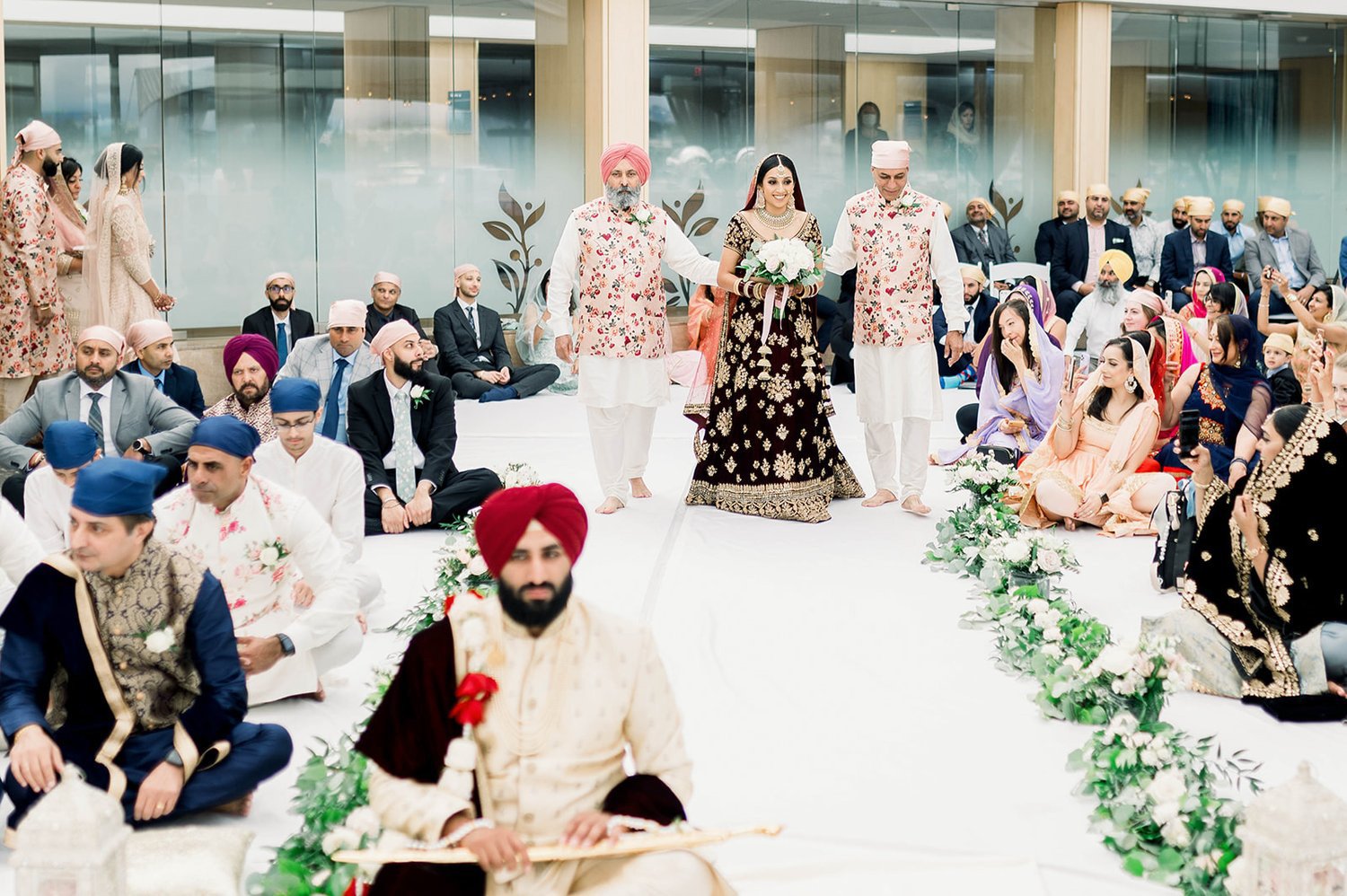 A bride in a red lehenga with gold accents is walked down the aisle by her parents as her groom awaits her
