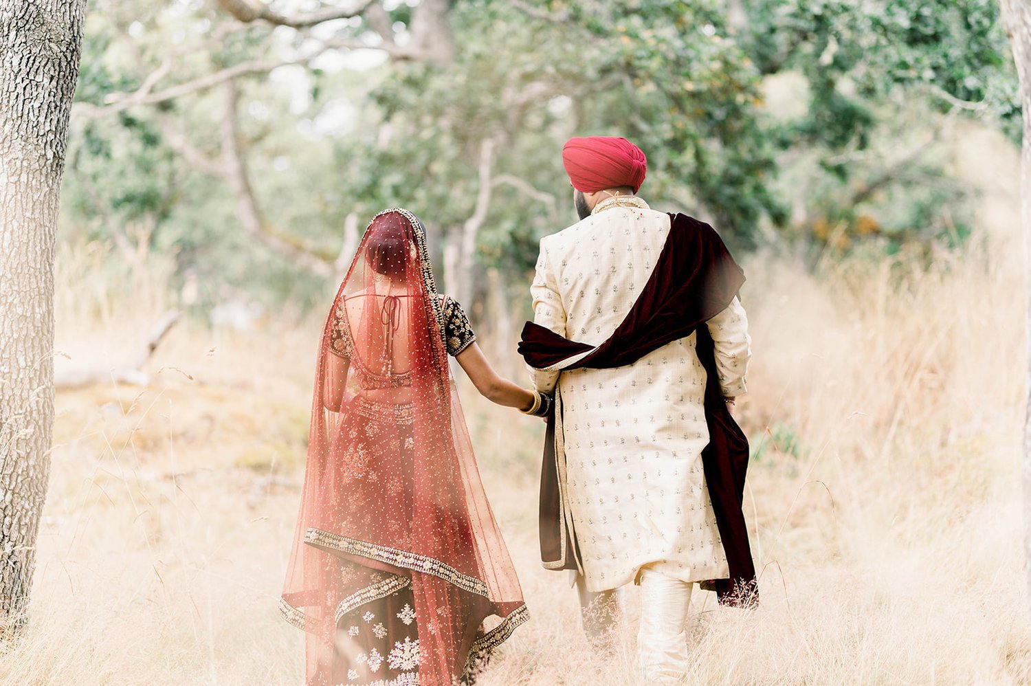 An traditional Indian couple walk away from the camera through romantic, dried, tall grass in Victoria BC 