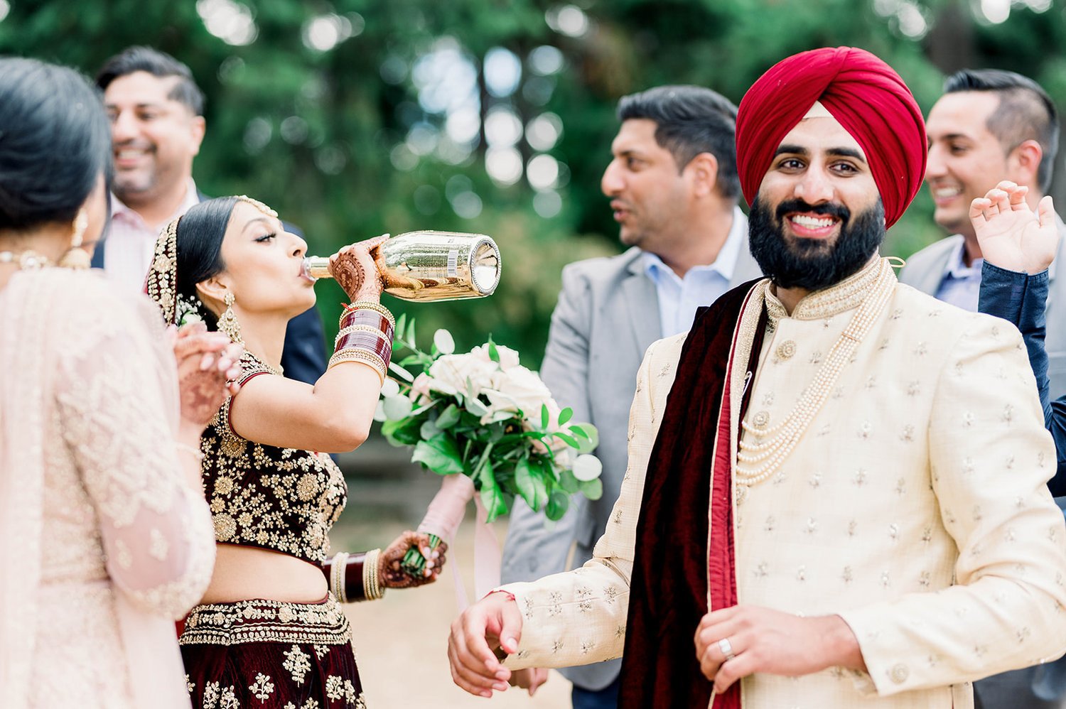 An indian groom smiles as his bride takes a swig of champagne from the bottle 