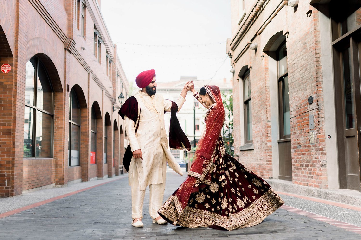 A groom twirls his bride on a historic street lined with brick buildings in Victoria BC. 