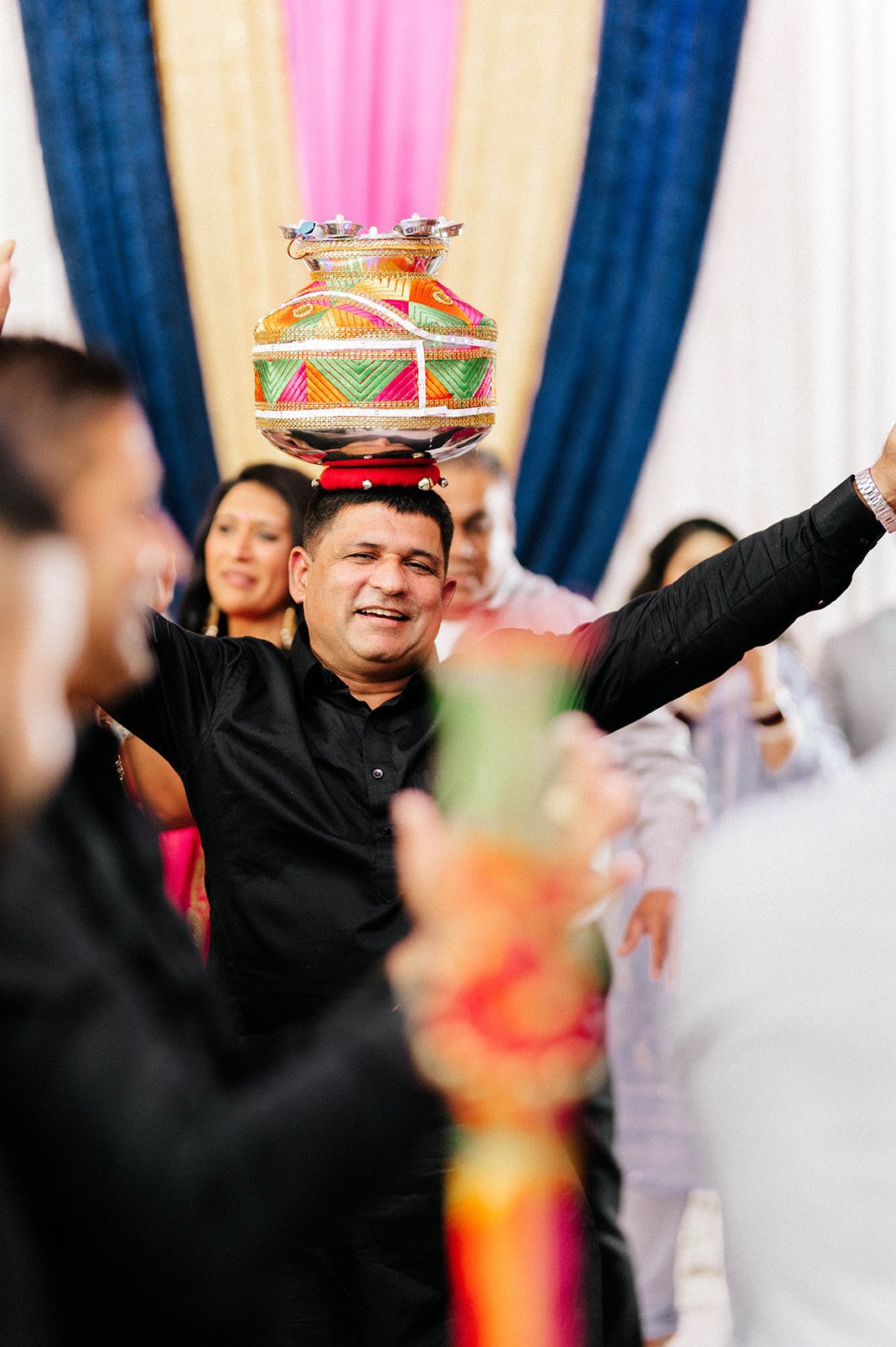 An indian in a black tunic dances with an ornamental jug balanced on his head 