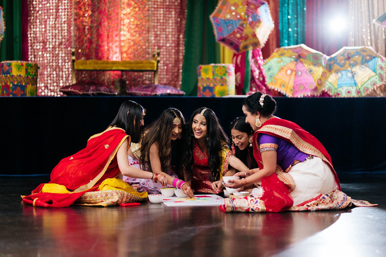 A group of women complete a rangoli board in front of vibrant colourful curtains.