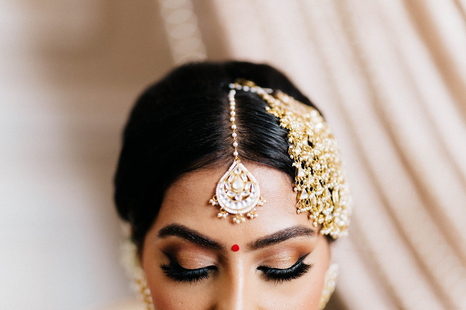 A South asian bride shows off her headpiece and eyeshadow as she looks to the ground in a close up photo. 