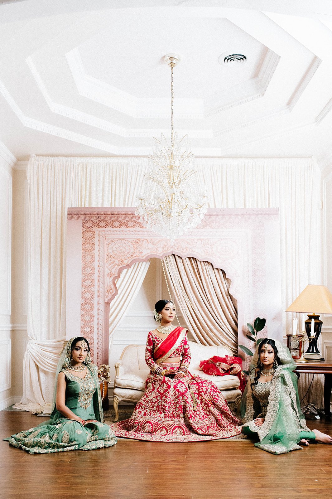 A indian bride poses beneath a chandelier in front of a backdrop with her two friends. 