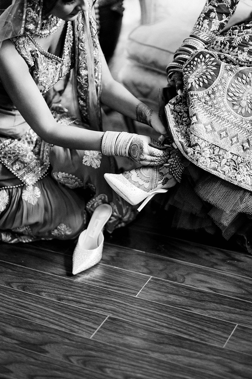 A close up black and white photo of a bride getting assistance putting her shoes on on her wedding morning. 