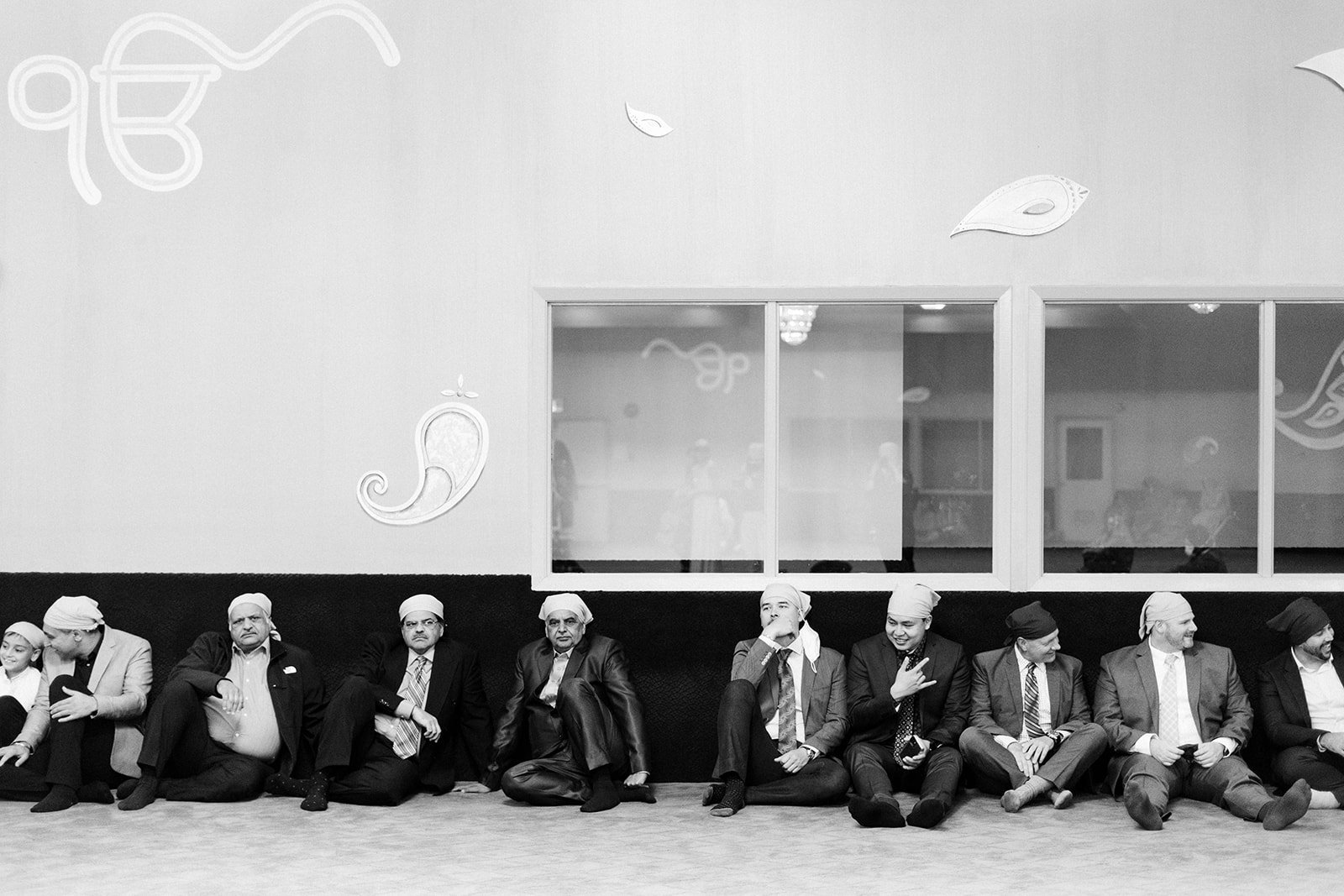 A line of 12 men sit cross legged along a wall 